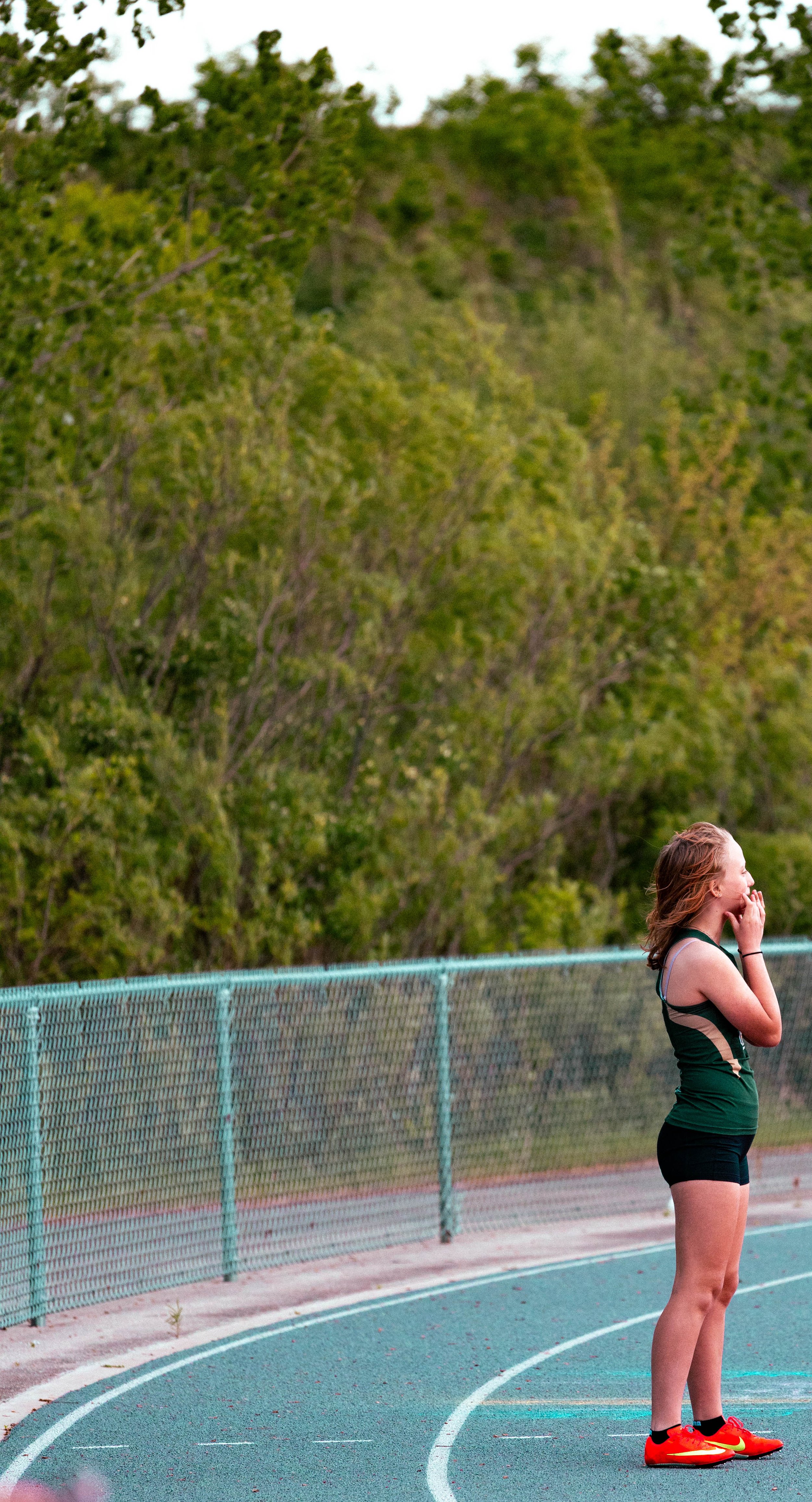 A girl standing on a running track, wearing a sports outfit and red athletic shoes, with her hands covering her mouth, near a green fence and lush trees in the background.