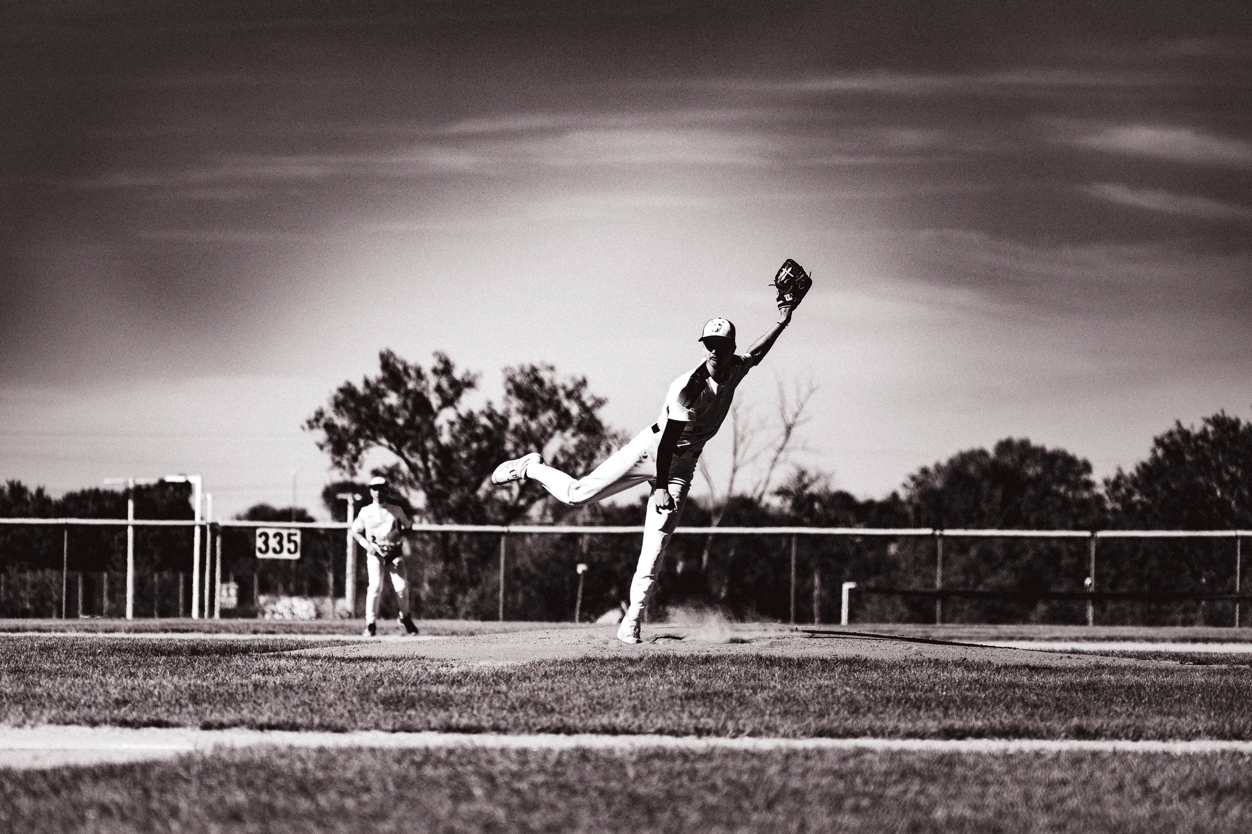 Black and white photo of a baseball player making a diving catch during a game, with other players in the background on the field and trees beyond the fence.