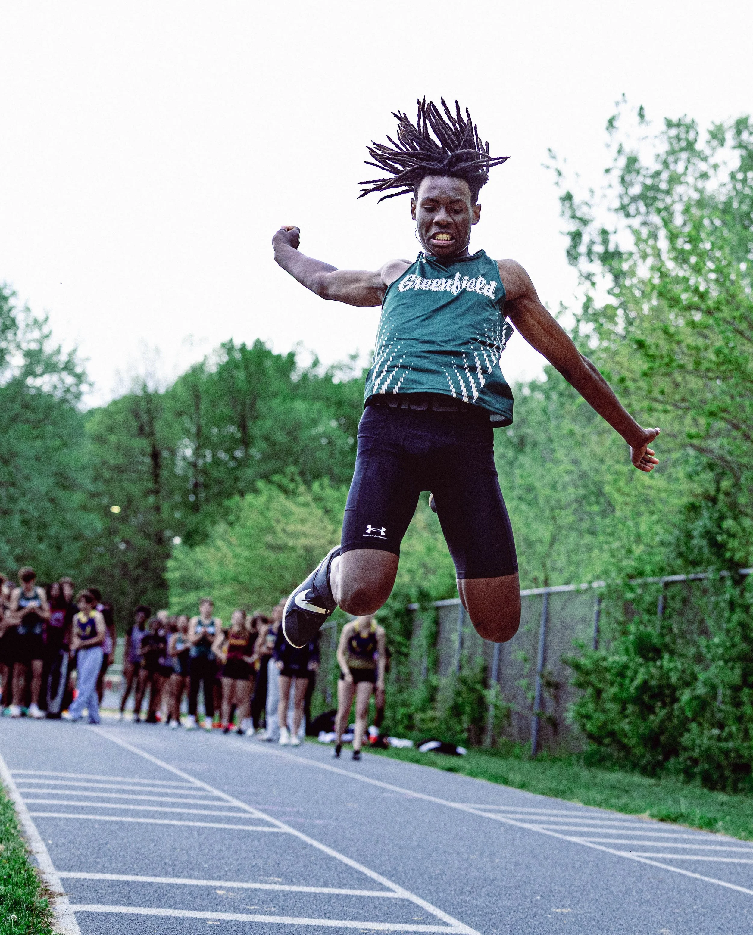A male long jumper in mid-air above a running track during a track and field event, with a group of athletes and spectators in the background and green trees surrounding the area.