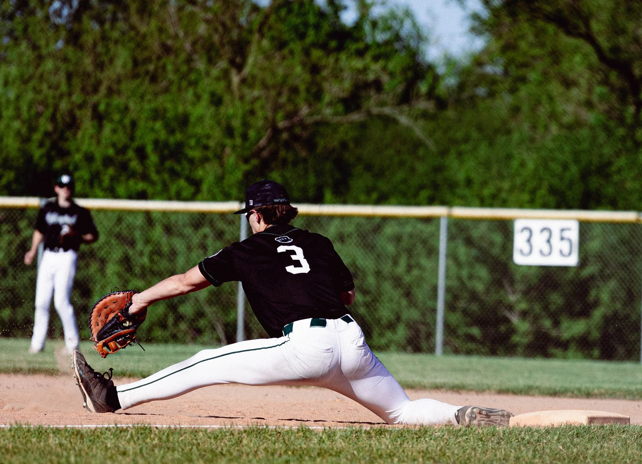 A baseball player diving to catch a ball in the field with a glove, while another player stands in the background near the fence.