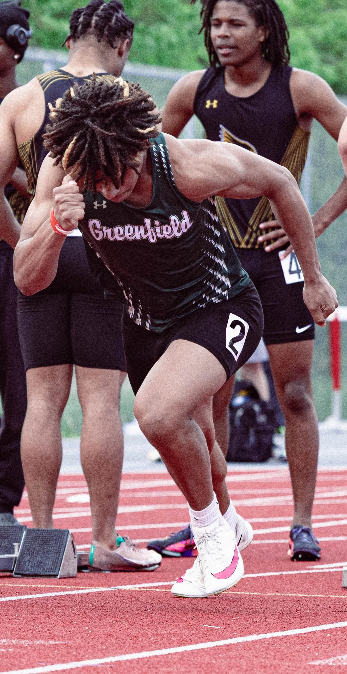A group of athletes at a track meet preparing for a race, with one runner in the foreground wearing a black uniform with 'Greenfield' and the number 2, stretching or warming up on a red running track.