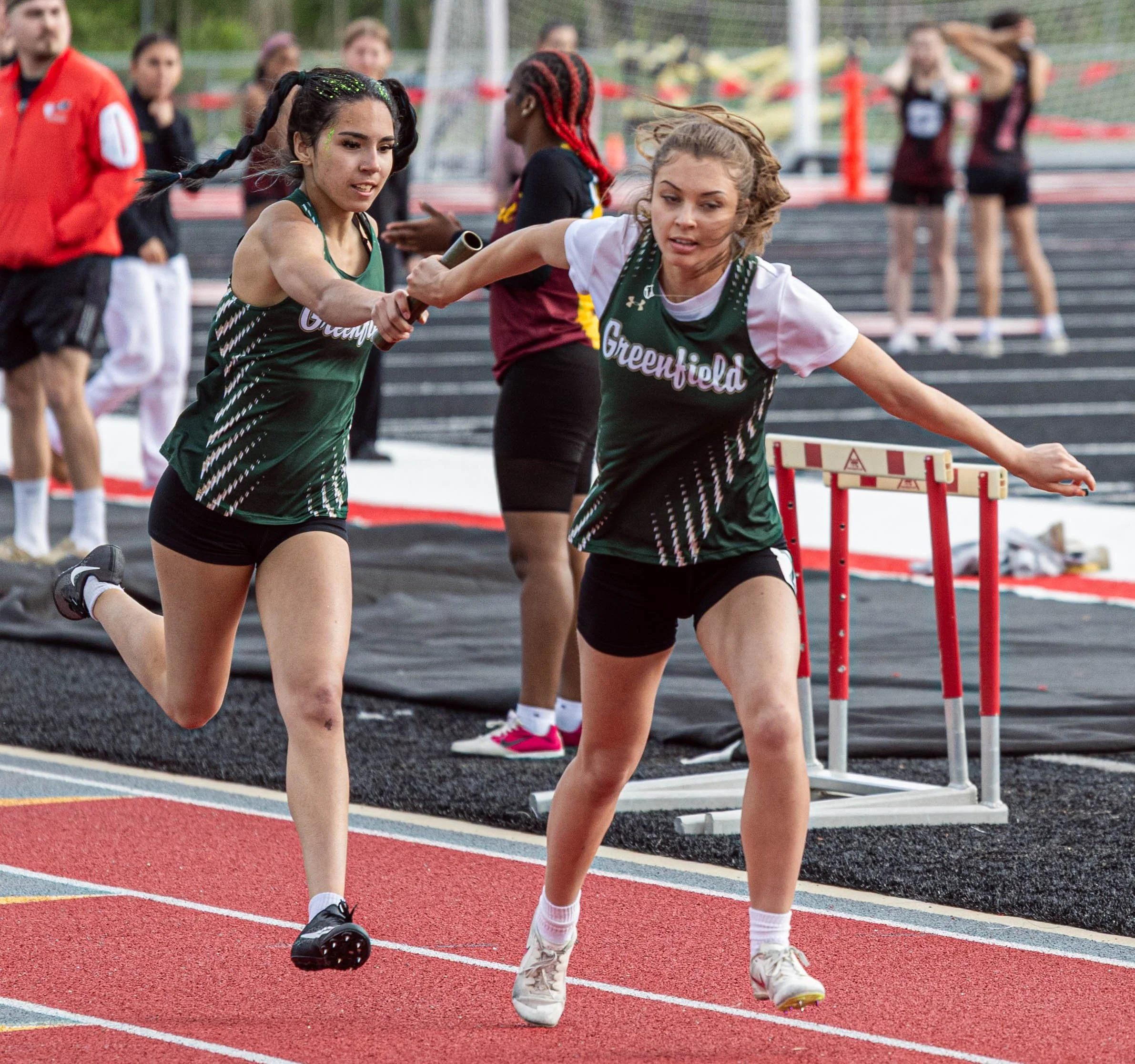 Two female athletes competing in a hurdle race on a track field, with spectators and officials in the background.