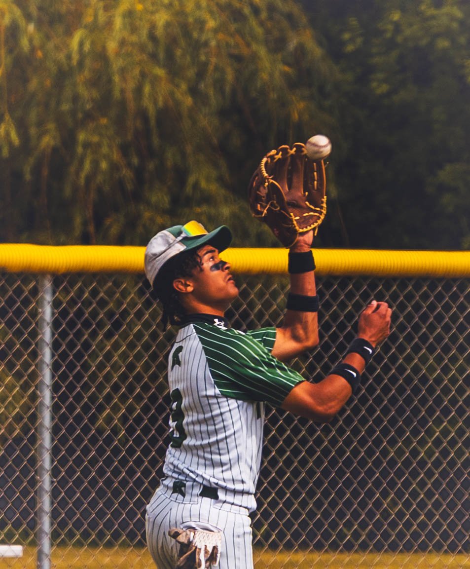 A baseball player in a striped uniform and cap catching a ball in his glove on a baseball field, with a chain-link fence and trees in the background.