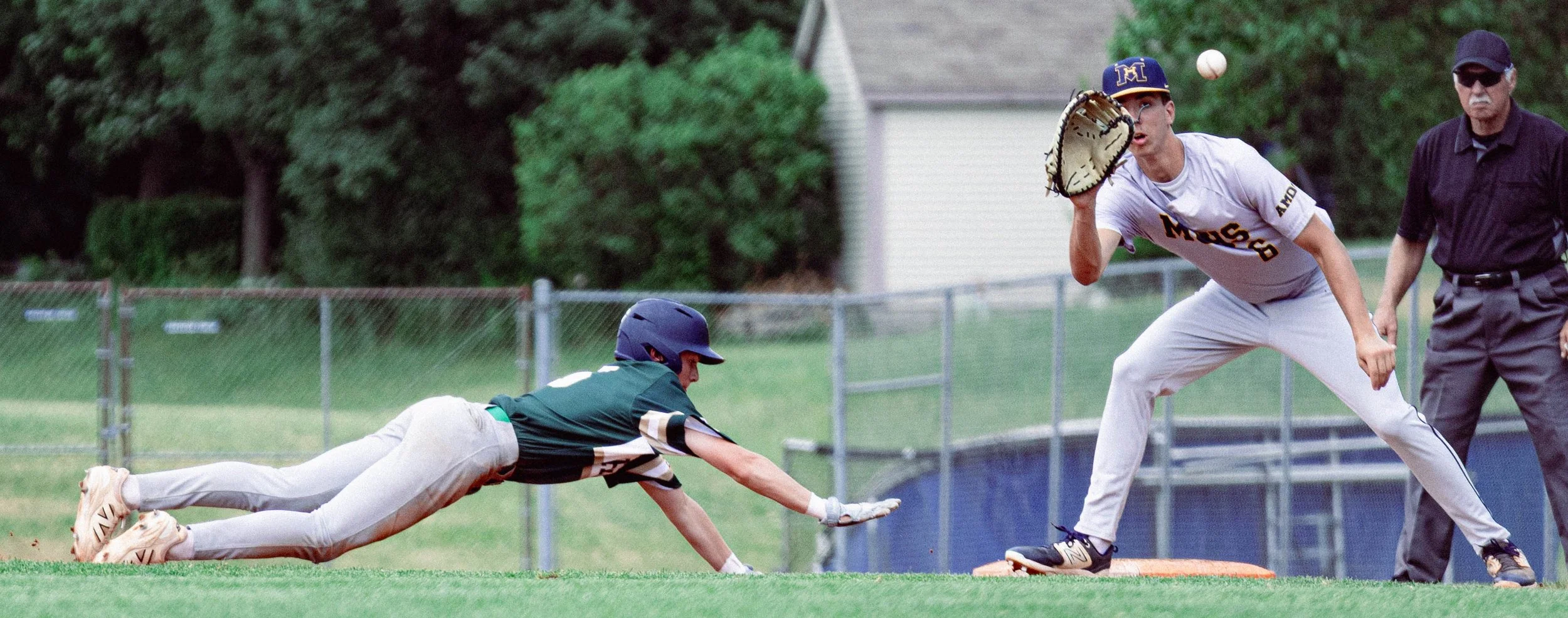 A baseball game with a player in a green uniform diving to catch the ball while another player in a white uniform and baseball mitt prepares to catch it, with an umpire observing nearby.
