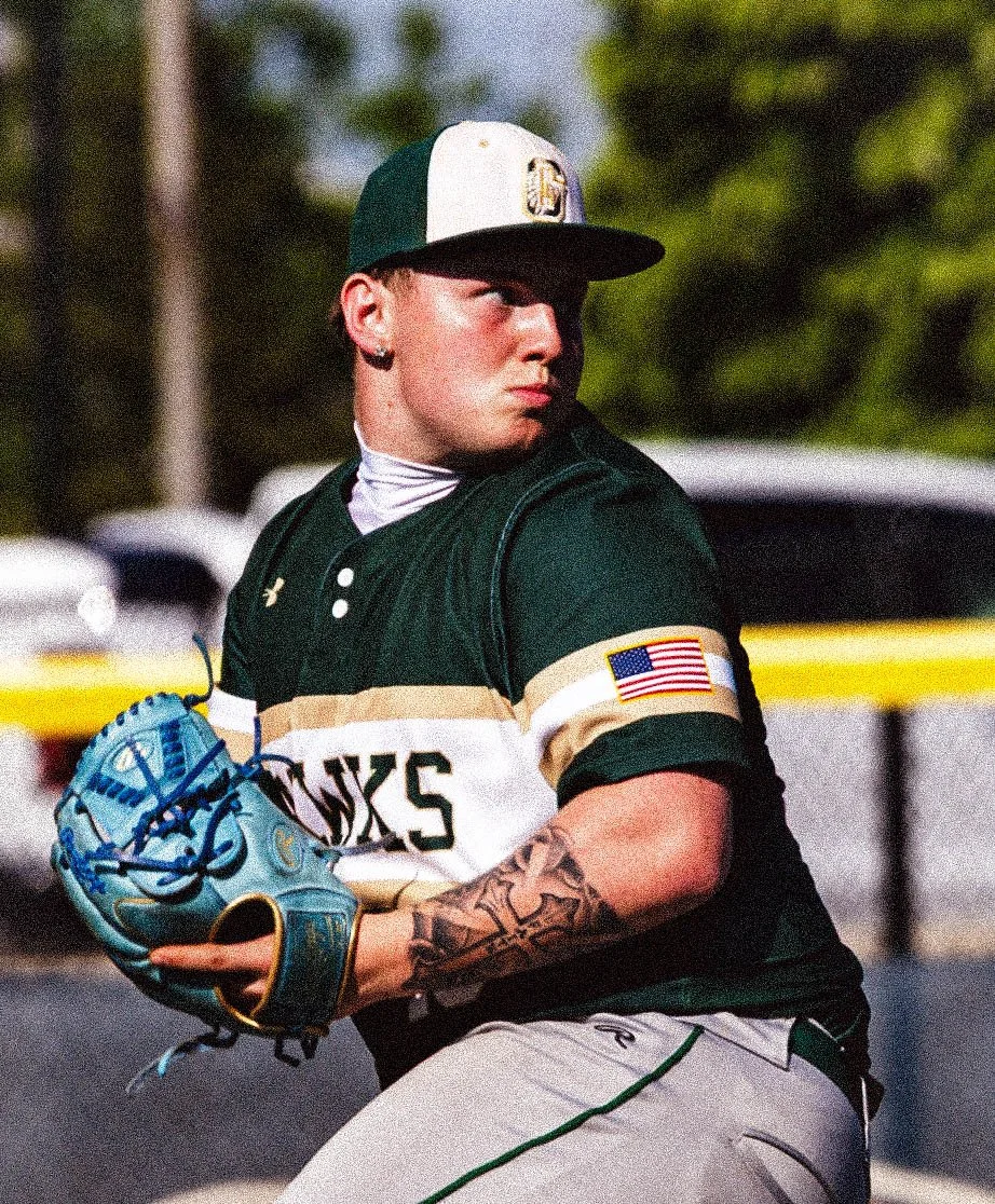 A young baseball player wearing a green and white uniform with an American flag patch on the sleeve, holding a blue baseball glove during a game.