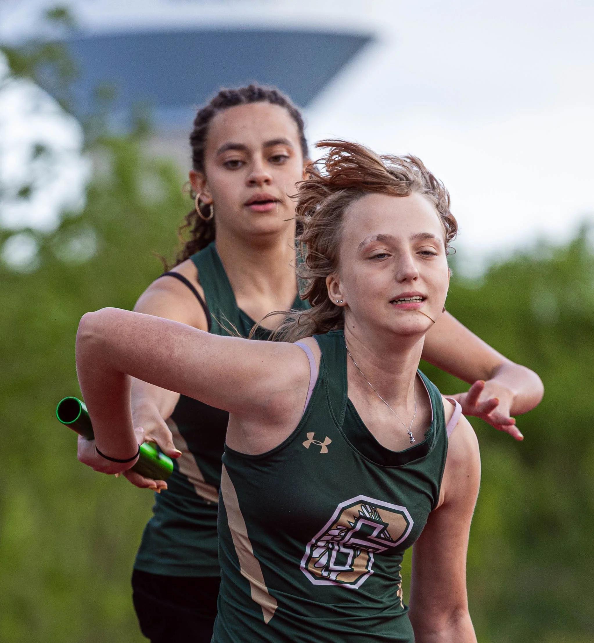 Two young women running together outdoors, wearing athletic wear, with one person holding a green baton in a relay race.