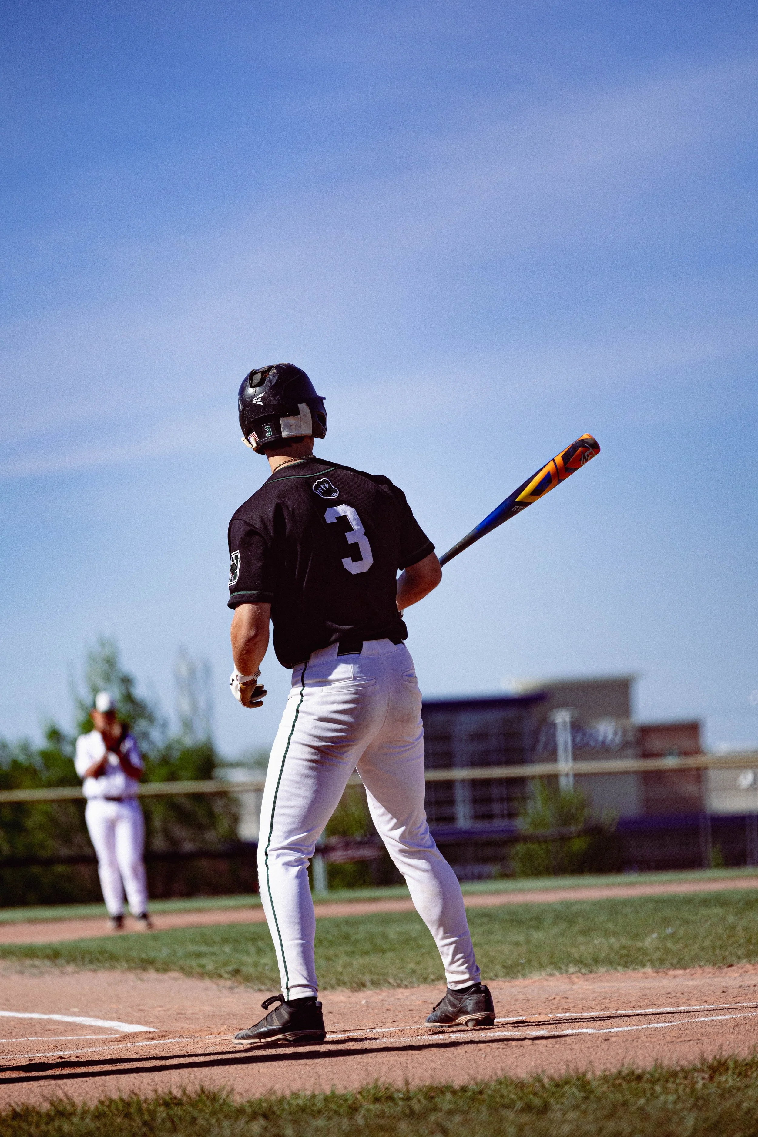 A baseball player wearing a black jersey with the number 3 on the back, white pants, black cleats, and a black helmet, holding a bat on a baseball field. A referee and another player are visible in the background.