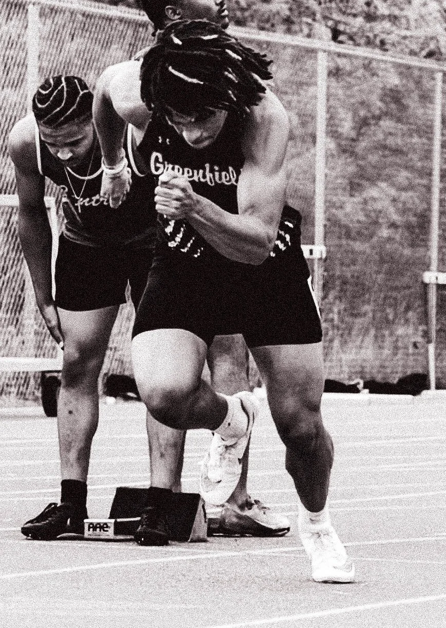 Athletes at the starting line in a track and field race. The runner in the foreground is wearing a sleeveless shirt and shorts, has dreadlocks, and is leaning forward in a sprinting position. The athlete behind is also wearing athletic gear and is pr
