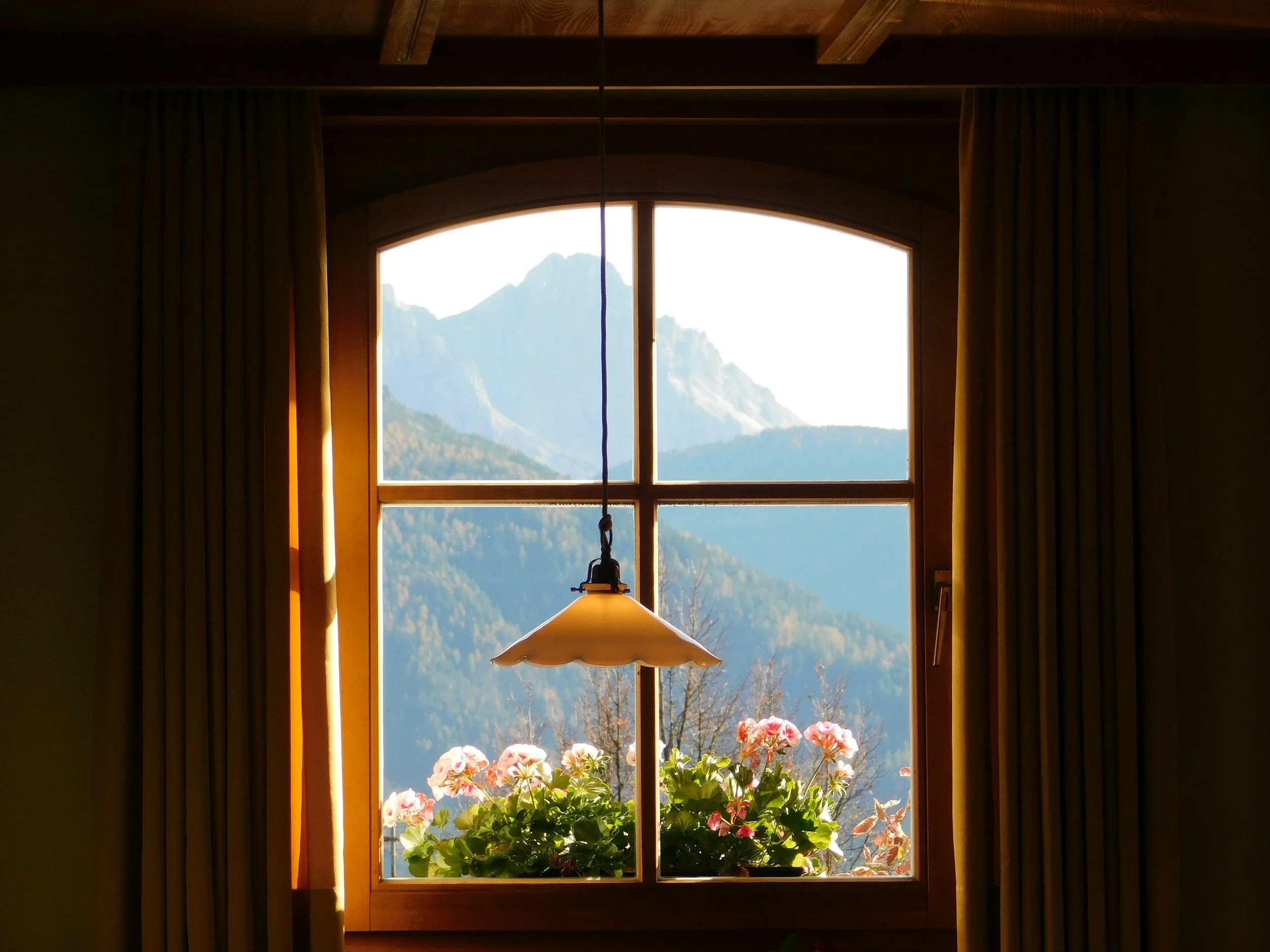 Inside view of a room looking out a large wooden-framed window with mountains in the background, flowers on window sill, and a hanging lamp in the foreground.