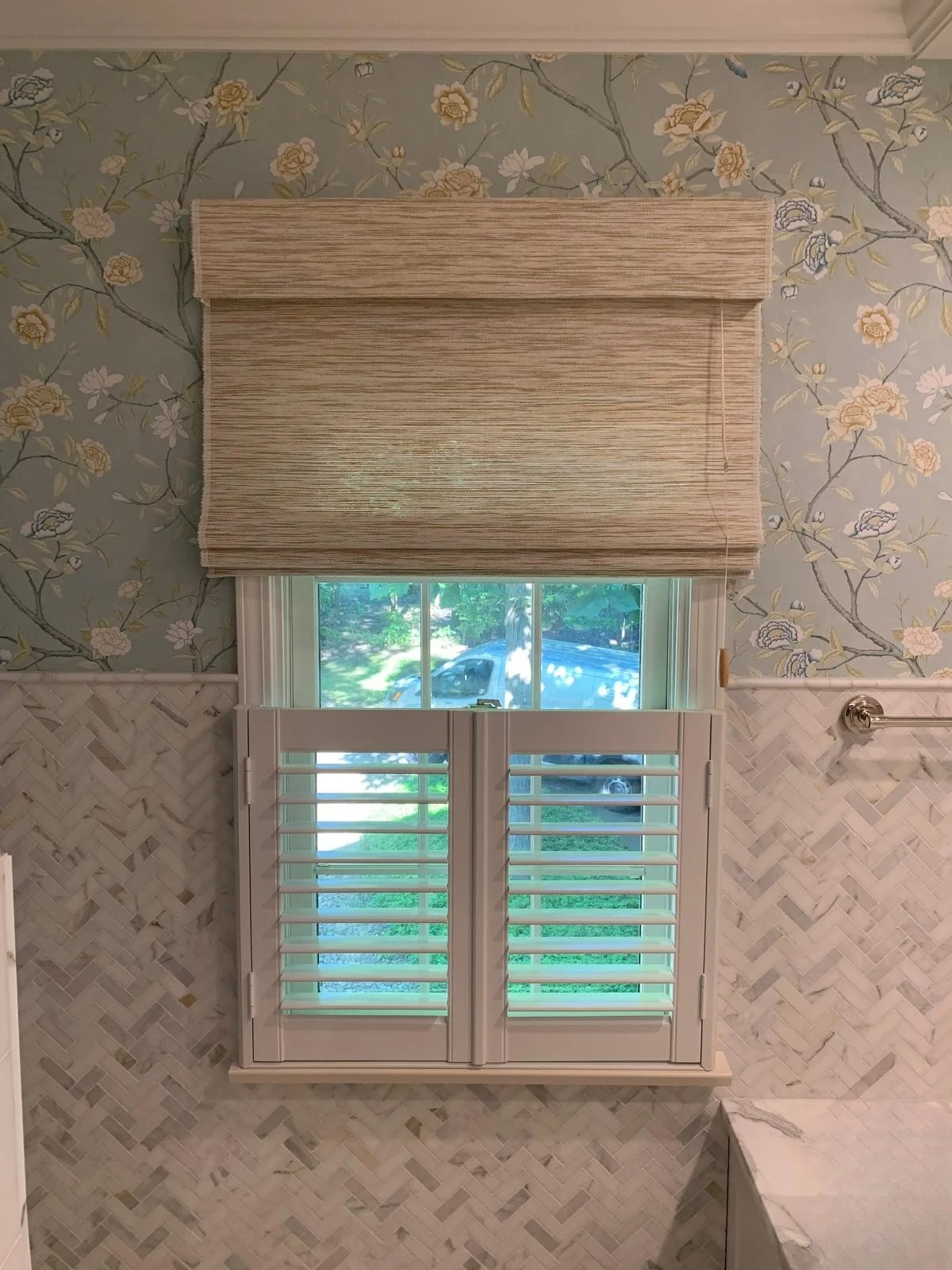 A window with white shutters and braided blinds above, in a room with floral wallpaper and a marble countertop.