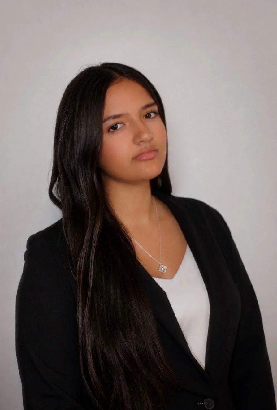 Young woman with long dark hair wearing a black blazer, white top, and a silver necklace, standing against a plain light gray background.
