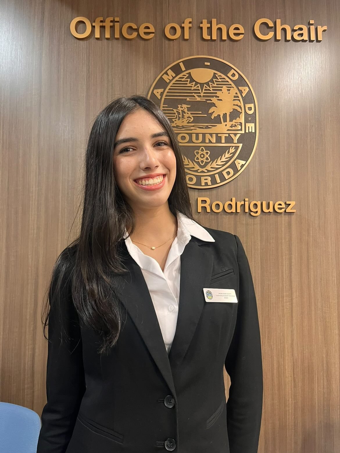 A young woman with long dark hair smiling, standing in front of a wooden wall with a gold seal of Miami-Dade County and the words 'Office of the Chair' and 'Rodriguez'.