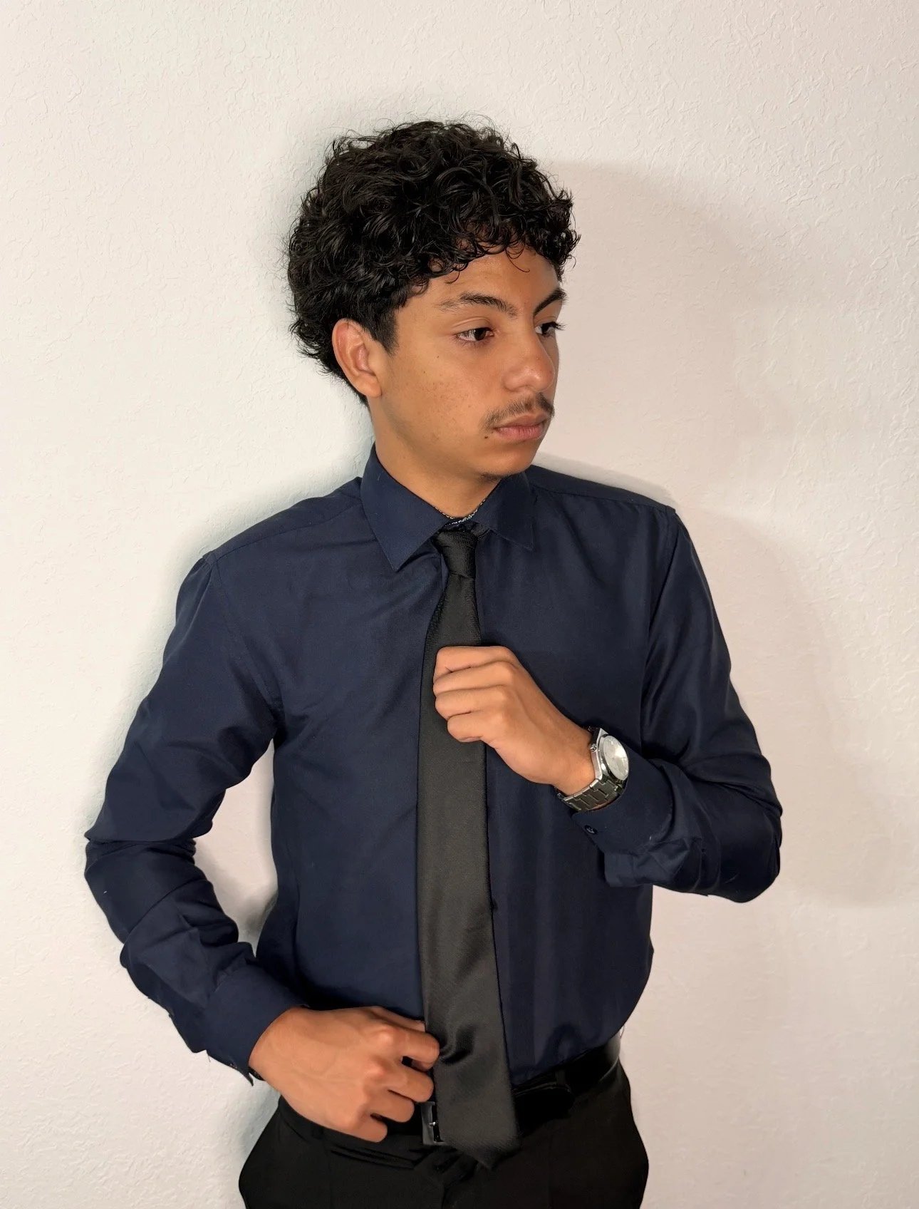 Young man with curly dark hair wearing a black dress shirt, black tie, and watch, standing against a white textured wall, adjusting his tie.