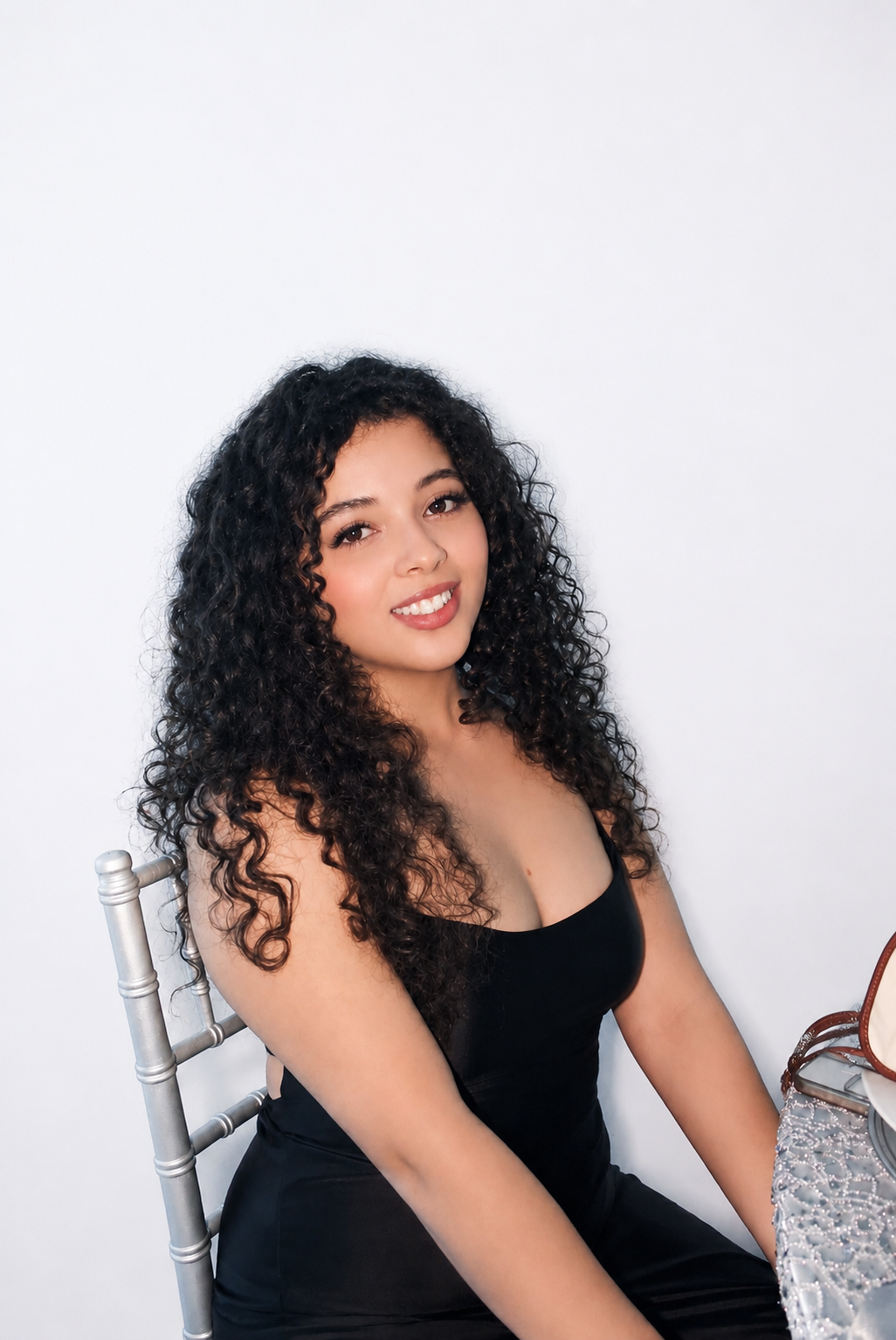 A young woman with curly dark hair wearing a black strapless dress, sitting in front of a plain white background, smiling at the camera.