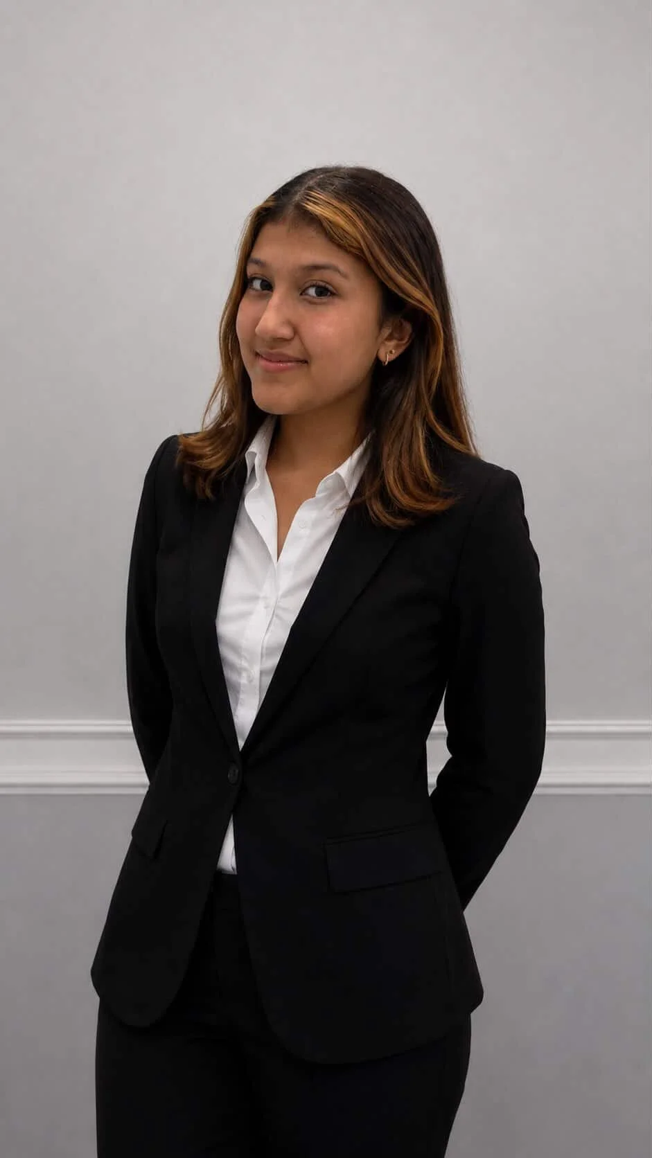 A woman in a black blazer and white shirt, standing against a plain gray wall.
