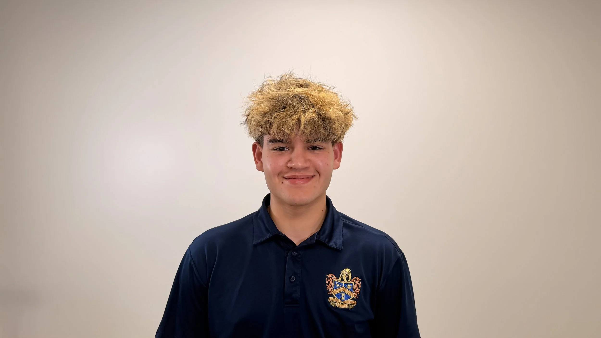 A young man with curly blond hair and fair skin, smiling softly, wearing a navy blue collared shirt with a crest embroidered on the chest, standing against a plain light beige background.