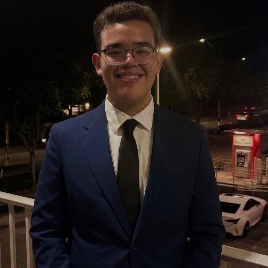 Young man in a blue suit, white shirt, black tie, and glasses smiling outdoors at night with cars and streetlights in the background.