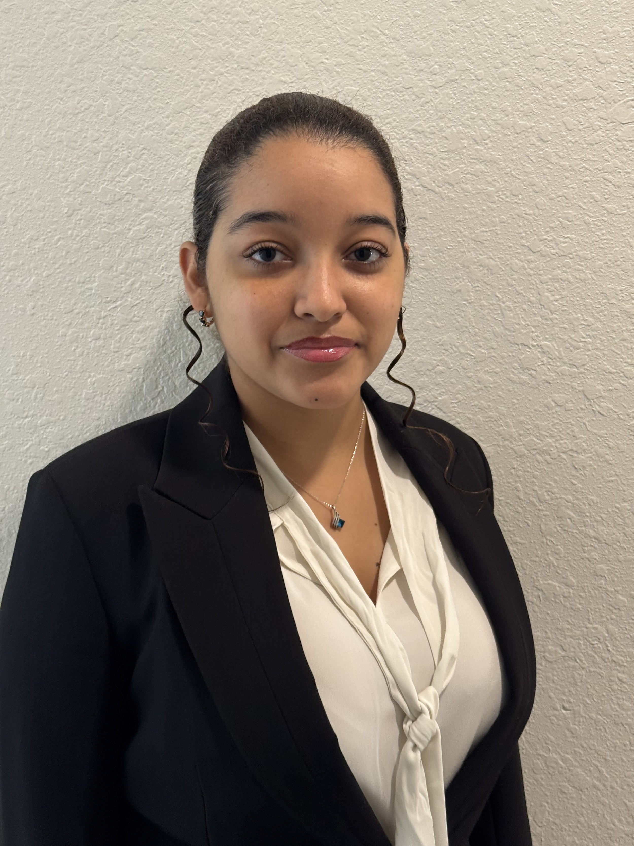 A young woman with dark hair styled in loose curls, wearing a black blazer and a cream-colored blouse with a tied knot, accessorized with earrings and a necklace, standing against a textured beige wall.