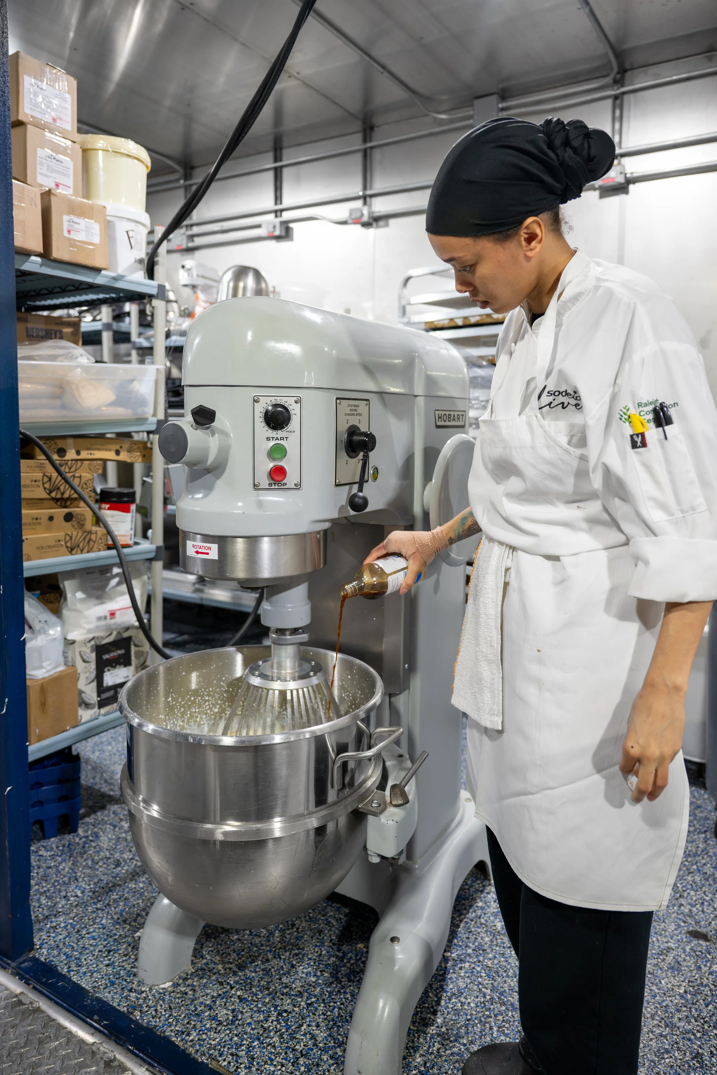 A person wearing a black headscarf and white chef's coat operating a large industrial kitchen mixer, pouring a liquid ingredient into the mixing bowl. Shelves with various boxes of supplies are visible in the background.