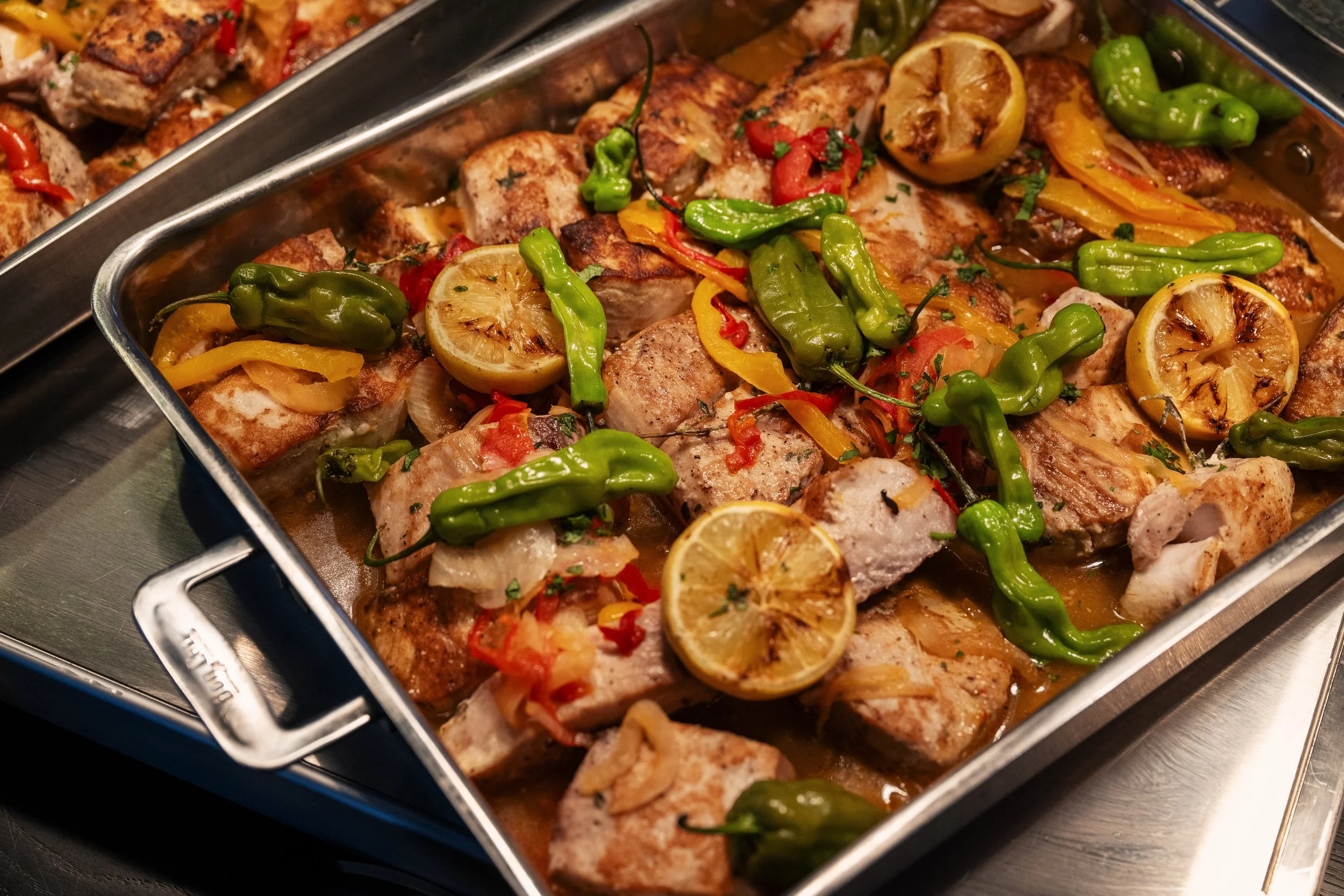A tray of cooked fish topped with grilled lemon slices, green and red chili peppers, and chopped herbs.