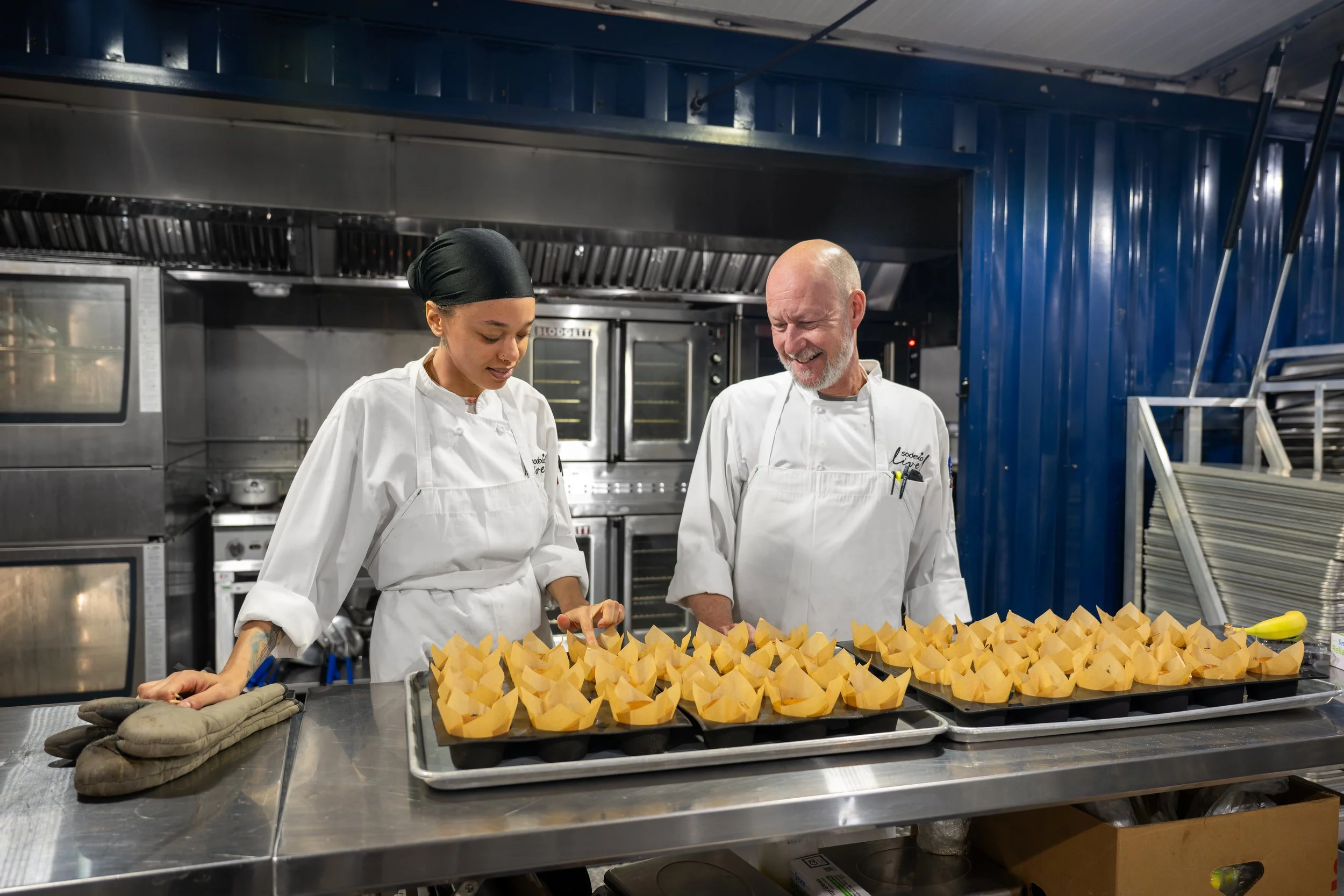 Two chefs in a professional kitchen preparing or decorating food on trays, with a row of yellow pastry wrappers on the counter.