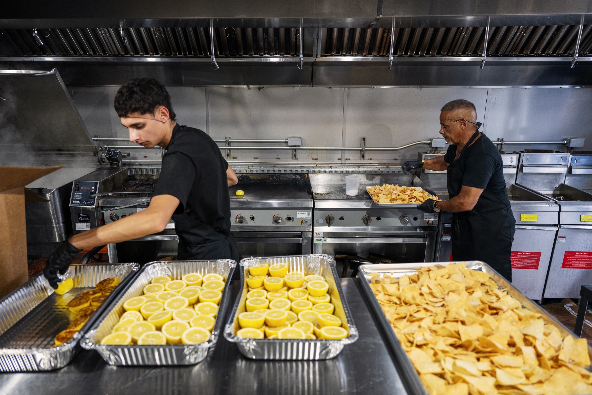 Two men working in a commercial kitchen preparing food with trays of sliced lemons and chips on the counter.