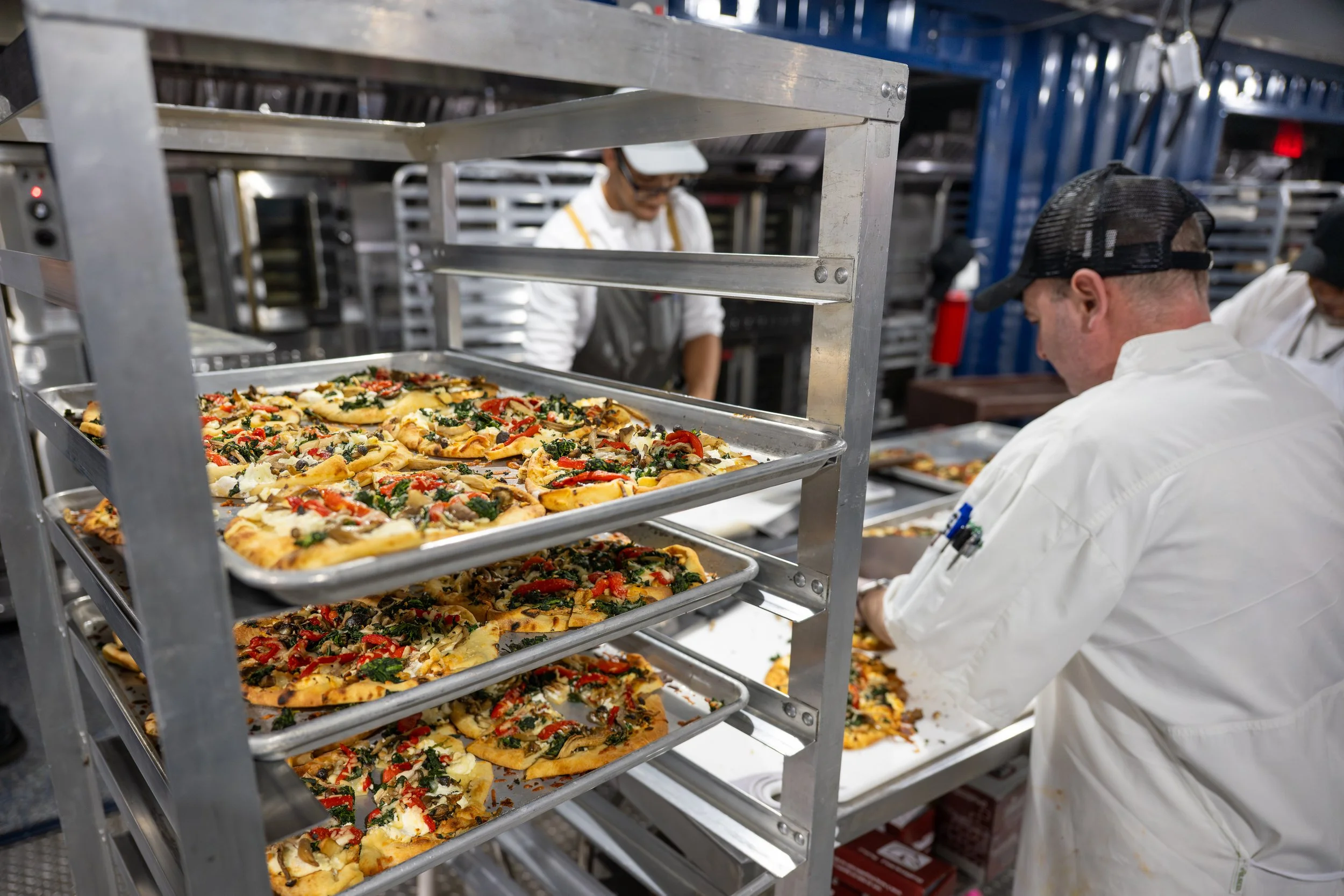 Chefs in a commercial kitchen preparing multiple trays of vegetable pizza.