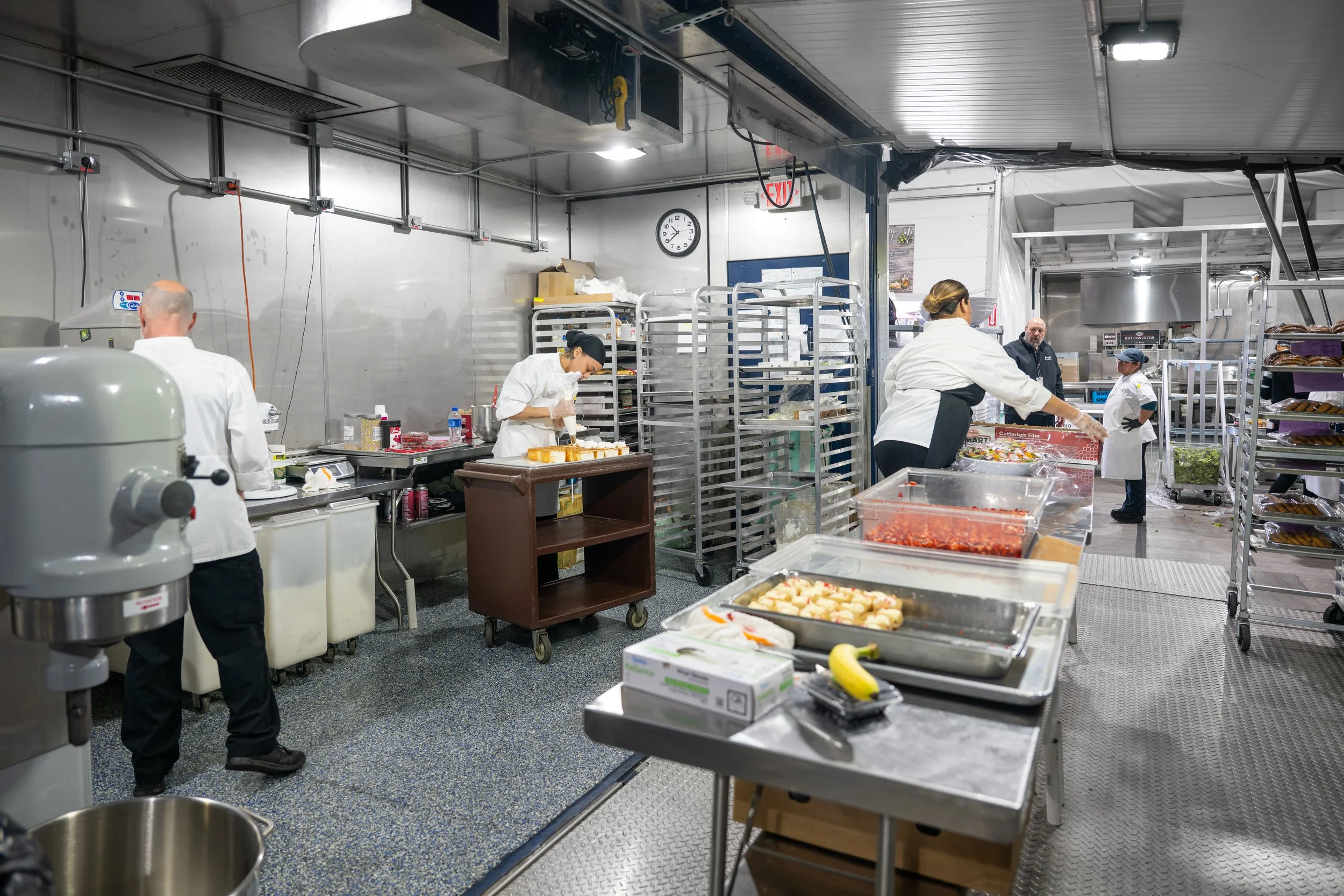 Commercial kitchen with employees preparing food, trays with baked goods, and various kitchen equipment.