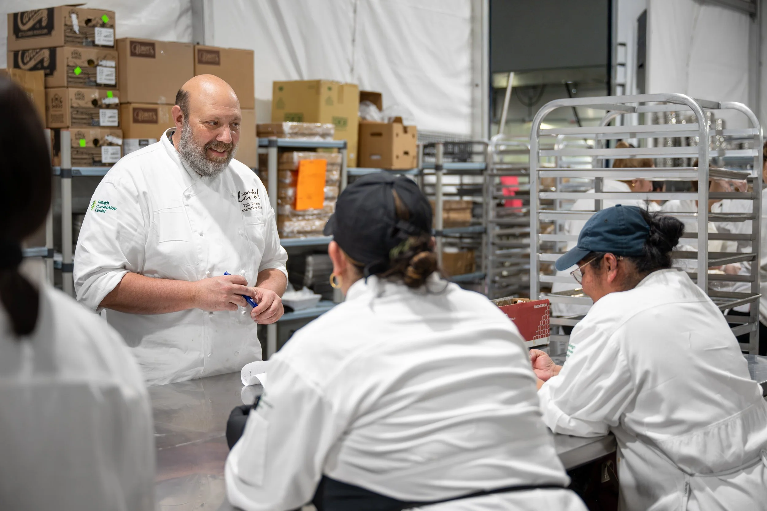 A group of people in white coats gathered around a table in a storage or packaging area, listening to a man with a chef who is speaking and holding a pen. Shelves with cardboard boxes are visible in the background.