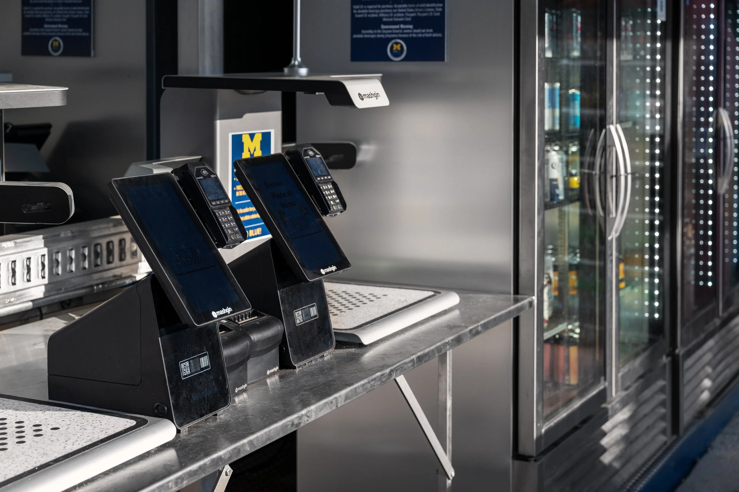 A row of three black and blue card payment terminals on a counter, with a sign behind them displaying the University of Michigan logo, and a glass-fronted refrigerator in the background.
