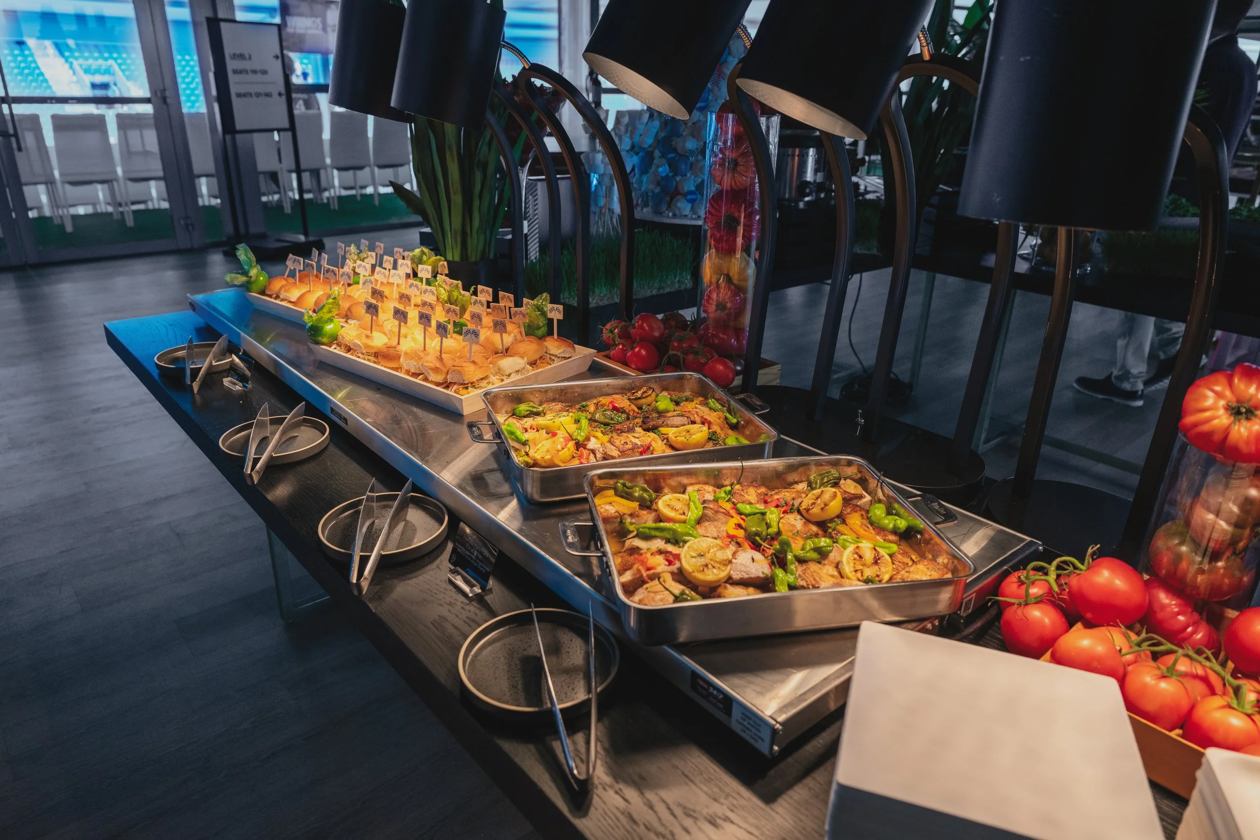 Buffet table with trays of cooked chicken and vegetables, tray of sliders with small flags, and bins of fresh tomatoes and peppers at a restaurant or event.