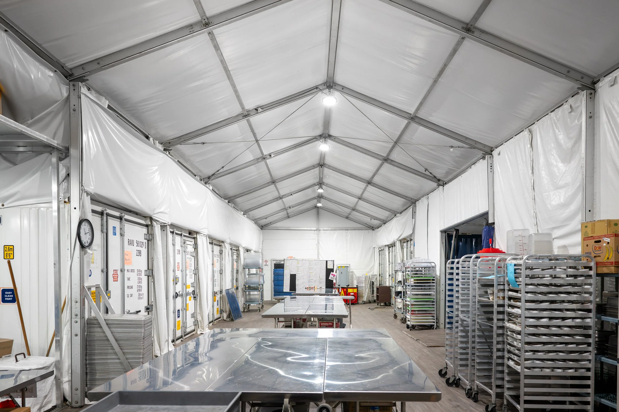 Interior of a large industrial or commercial food service tent, with metal tables, carts, and storage racks.