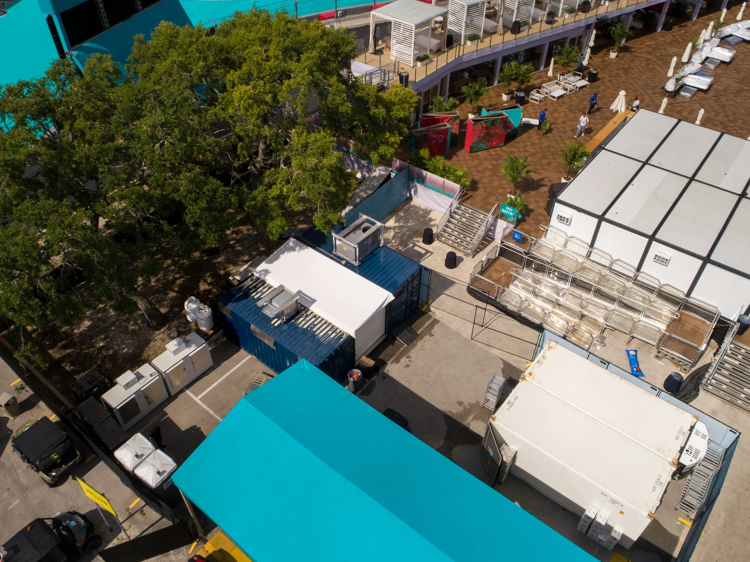 Aerial view of a mobile kitchen area near hospitality areas with lounge beds, umbrellas, greenery, and people walking around.