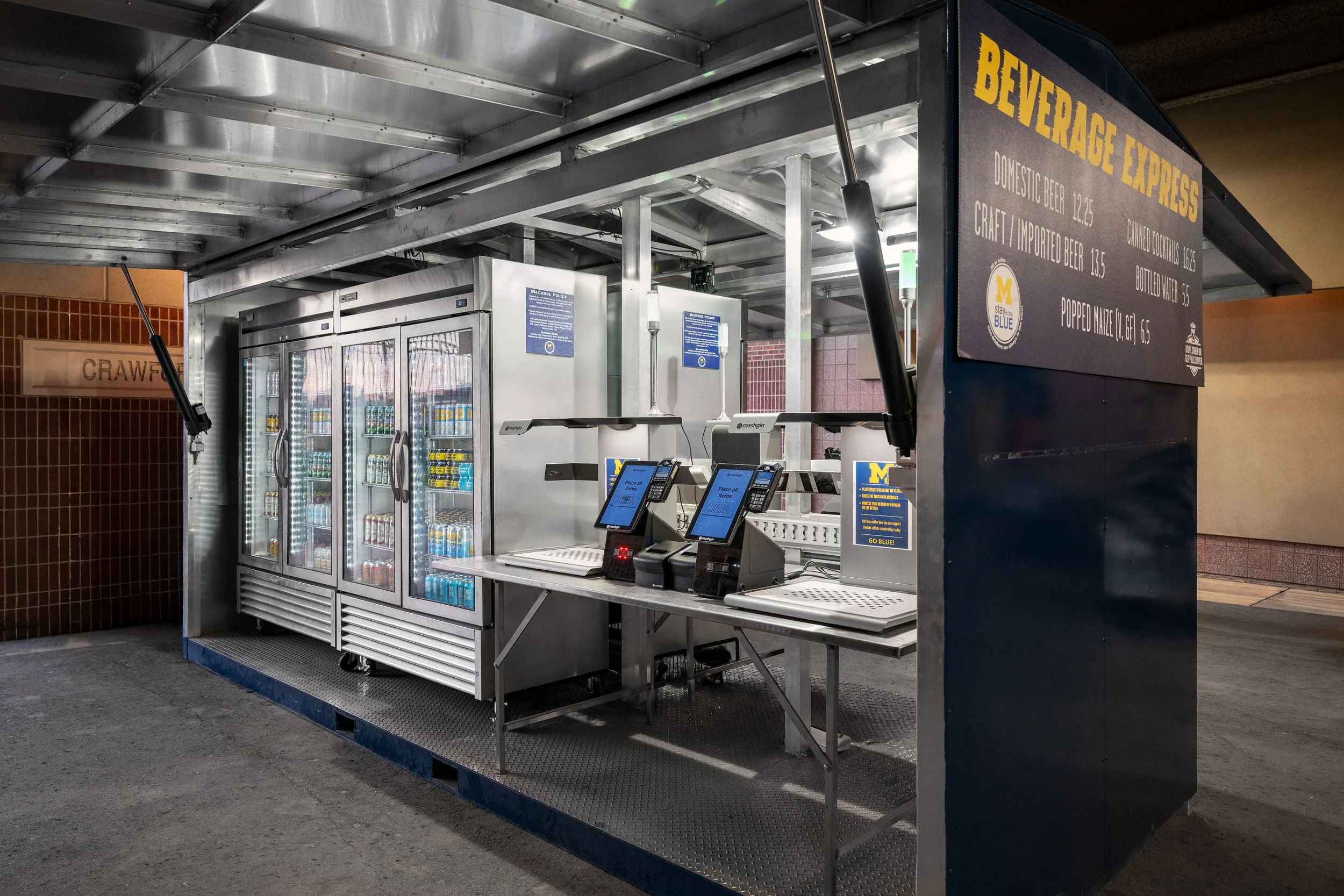 A beverage stand with a menu sign selling domestic and craft beer, cocktails, water, and popcorn, with drink vending machines and payment terminals underneath a metallic canopy.