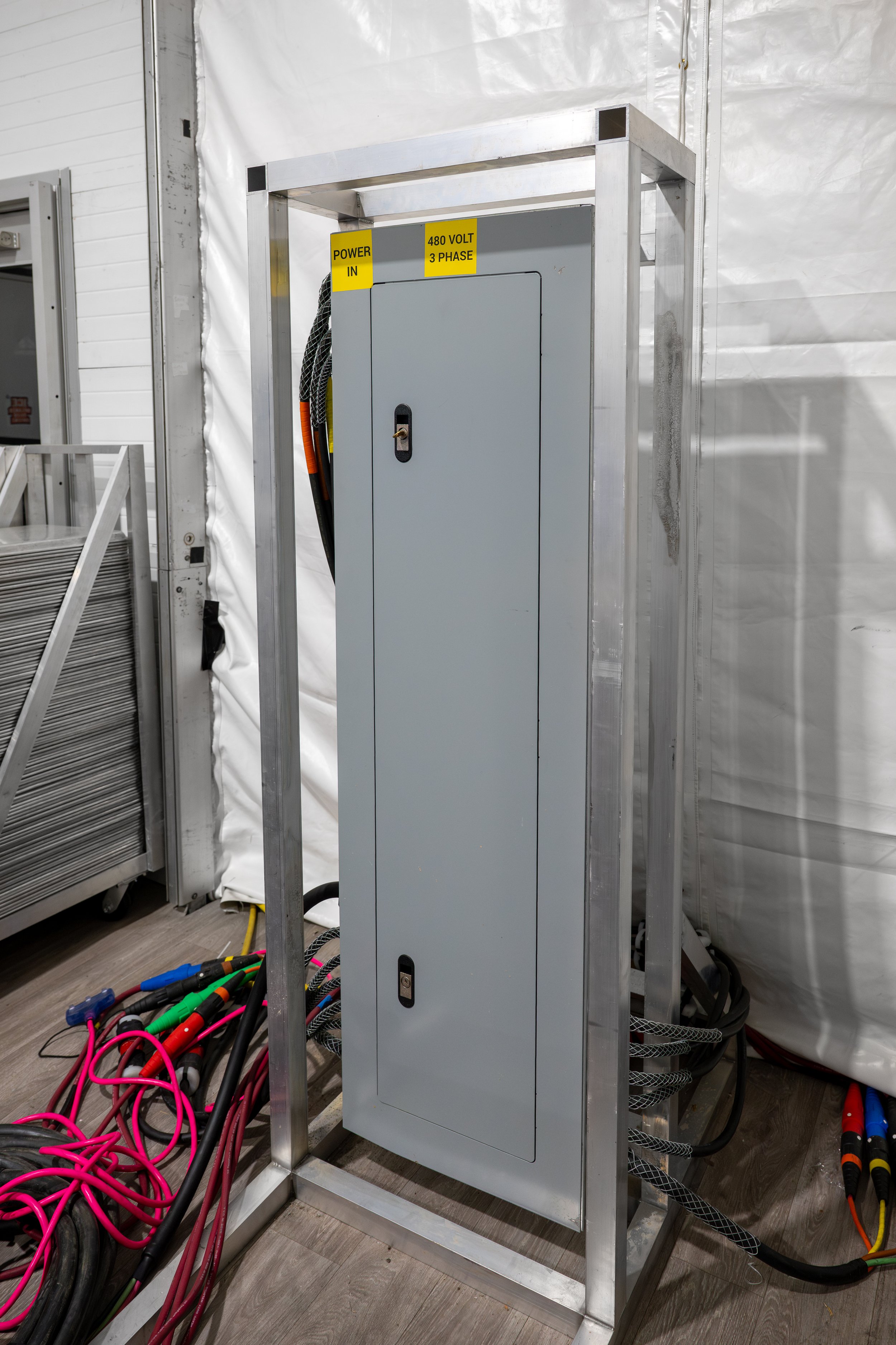 Electrical cabinet in a metal frame with multiple colorful cables on the floor, labeled signs indicating power input and voltage specifications.