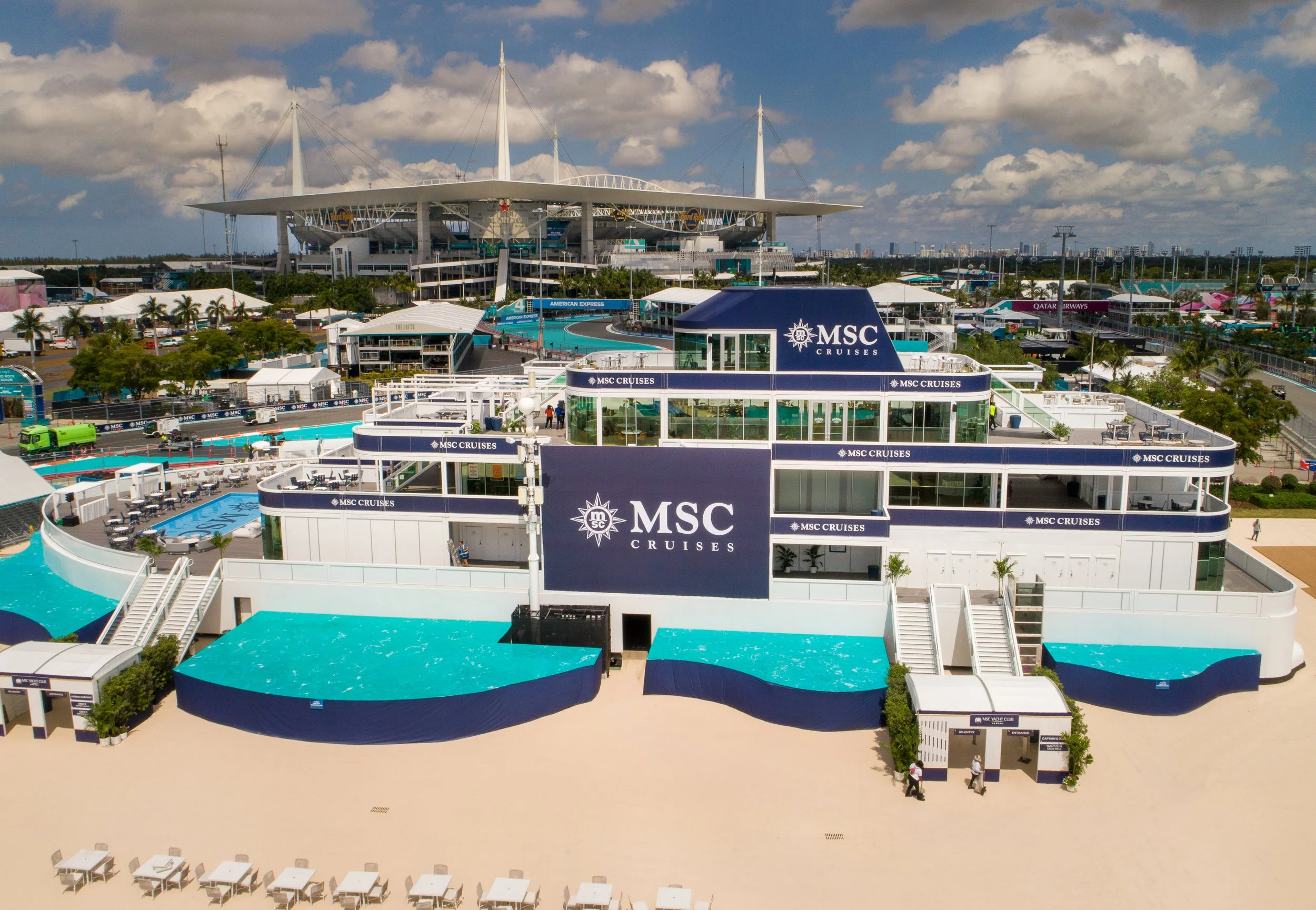 View of the MSC Cruises building with pool area at the Miami GP formula 1 race with a large stadium-like structure and partly clear sky in the background.