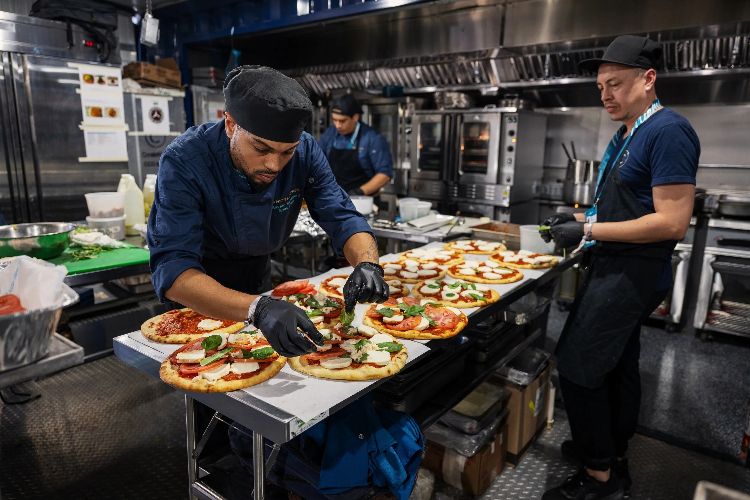 A commercial kitchen with three chefs preparing multiple pizzas on a tray. The pizzas have various toppings, including tomatoes, cheese, and greens.