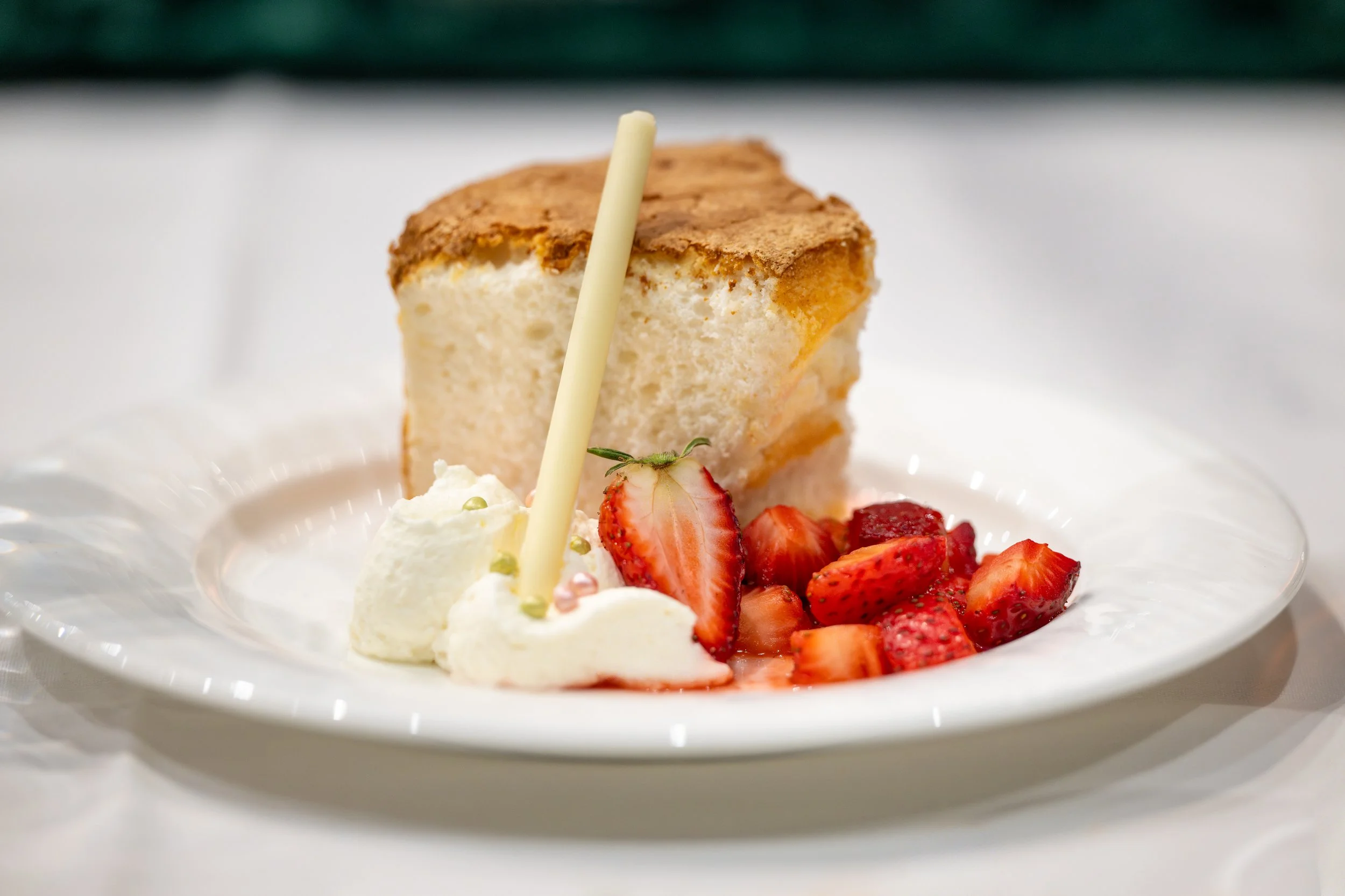 Slice of layered cake with frosting, served with fresh strawberries, whipped cream, and a white chocolate decoration on a white plate.
