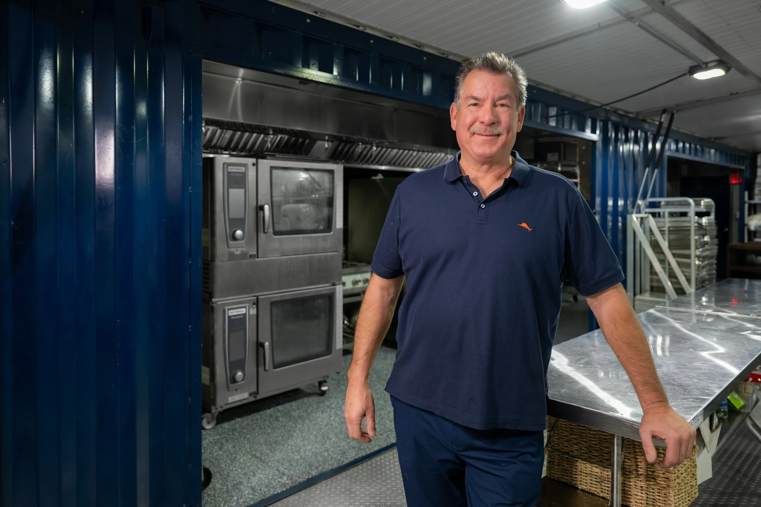 A middle-aged man, owner of MES John Hughes, in a dark blue polo shirt and matching pants standing inside a commercial kitchen or bakery, with stainless steel appliances and equipment in the background.