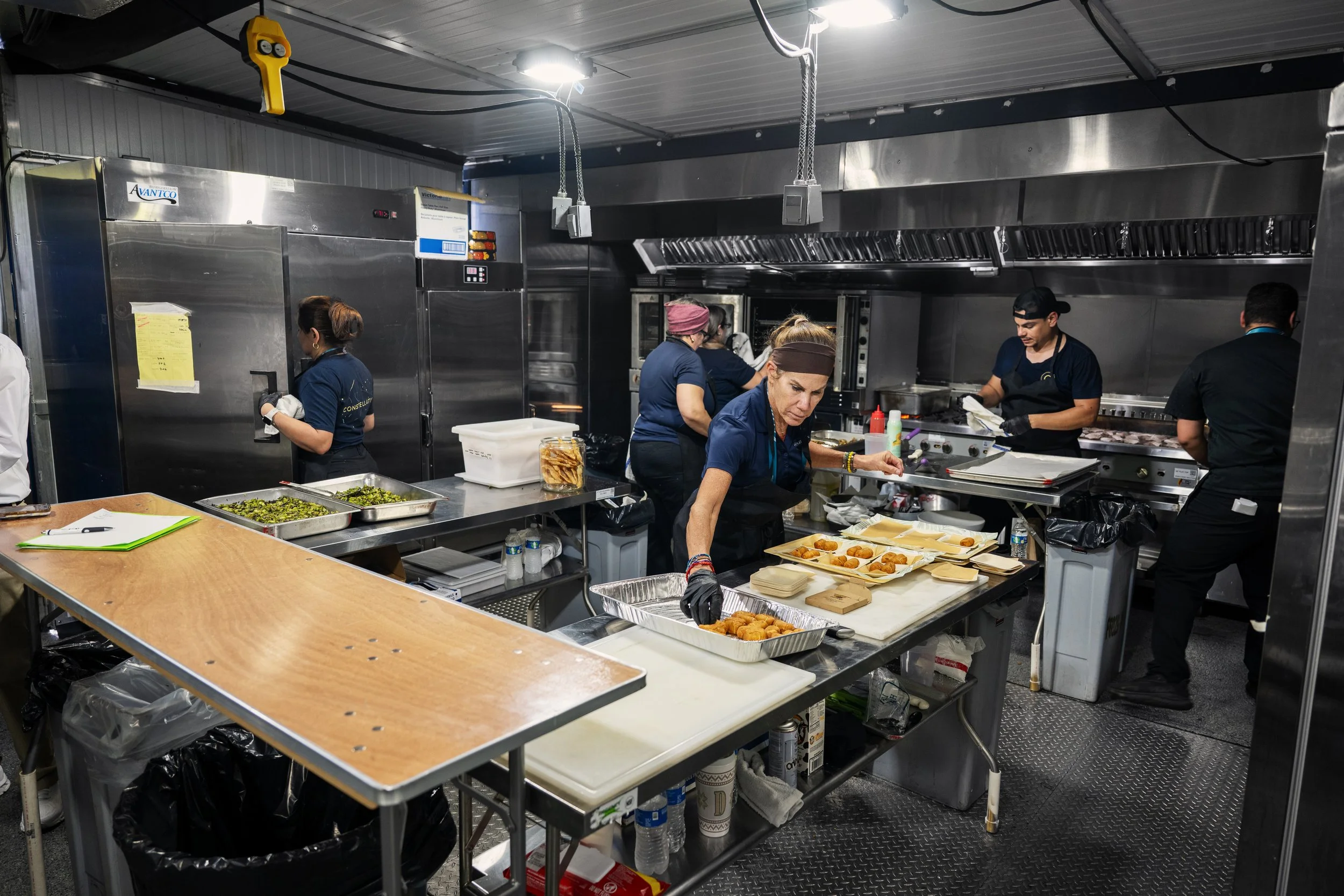 Kitchen with multiple workers preparing food, trays of food on the counter, industrial appliances, stainless steel surfaces, and various kitchen tools.