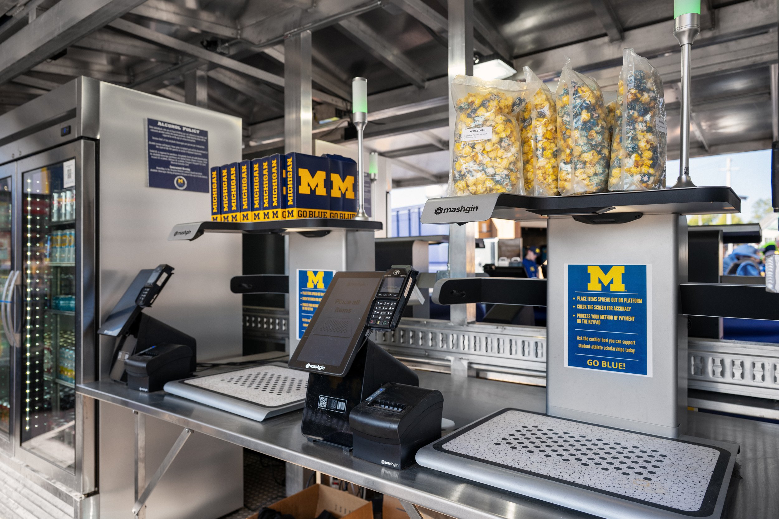 A concession stand checkout counter at the University of Michigan, with bags of popcorn, a cash register, and menus, under a metal ceiling with industrial lighting.