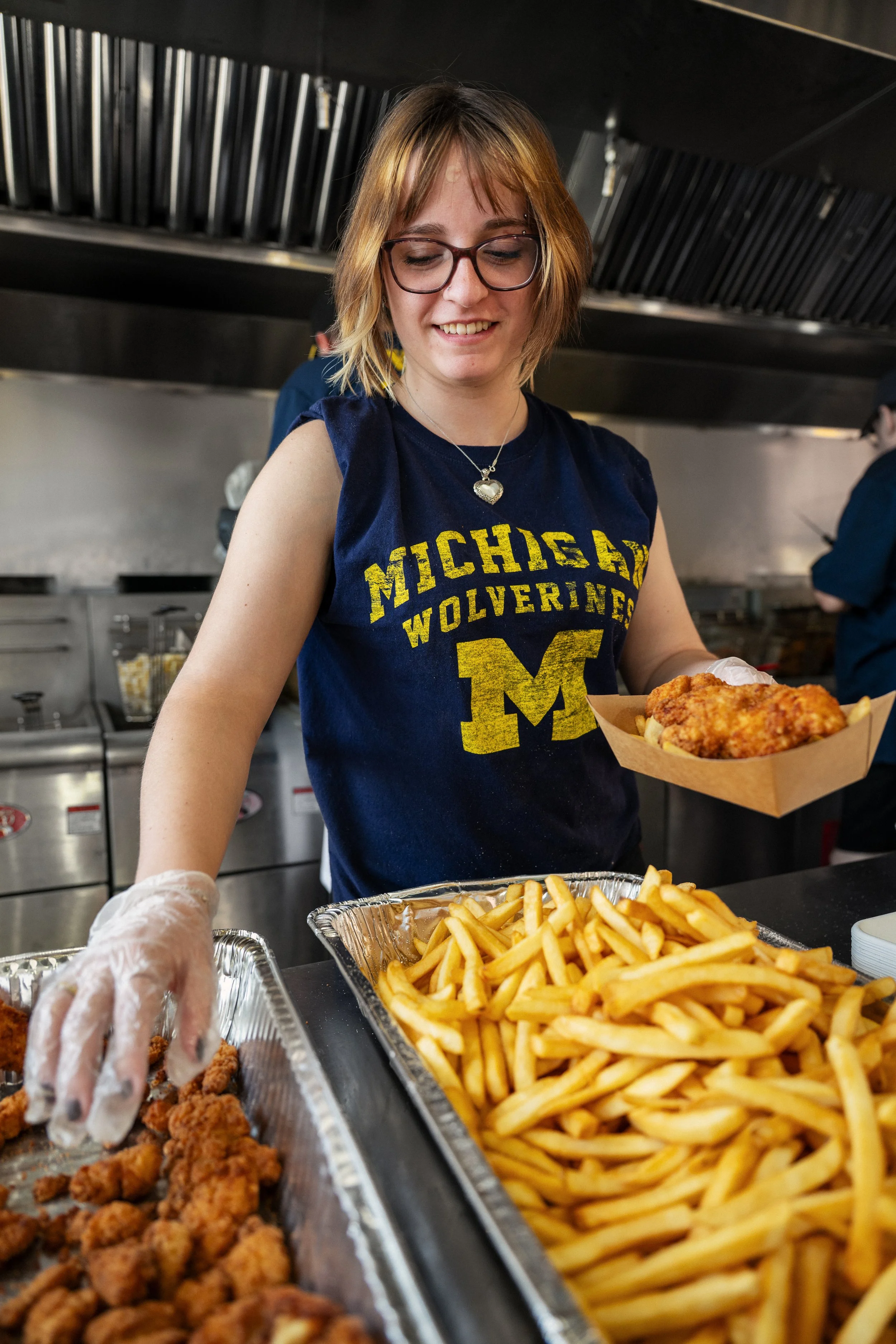 A woman wearing glasses and a Michigan Wolverines shirt serving French fries and fried chicken at a temporary mobile kitchen.