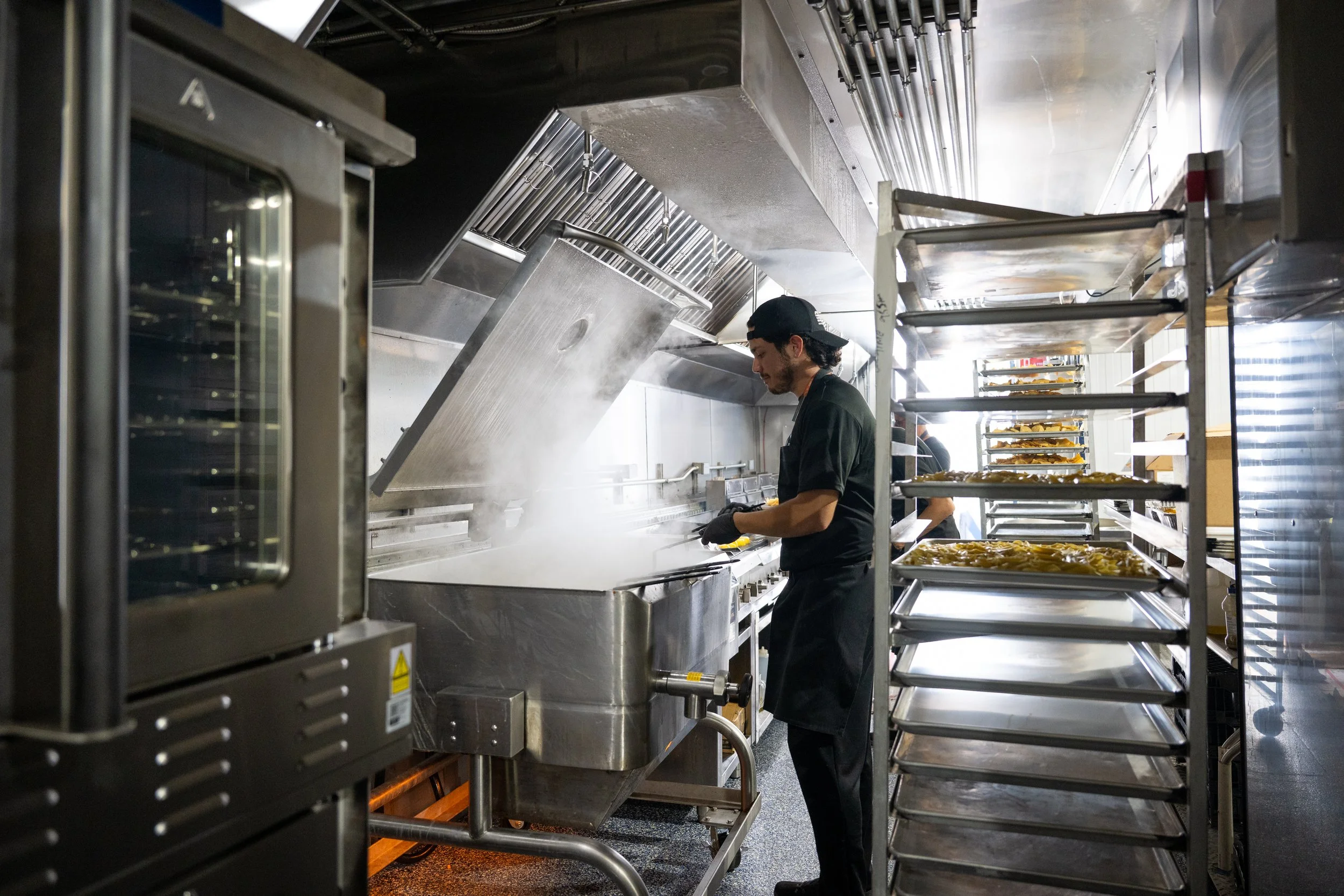 A man working in a commercial kitchen, standing next to stainless steel racks filled with trays of food. He is wearing a black shirt and a black cap, and appears to be preparing or cooking food.
