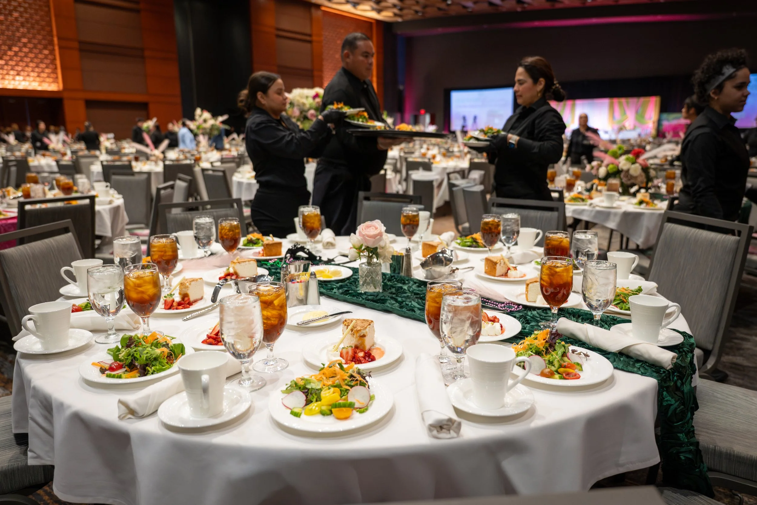 A banquet hall with a table set for dinner, featuring plates of salad, slices of cake, glasses of iced tea, cups, and utensils. In the background, waitstaff in black uniforms serve food, with a large screen and floral arrangements visible.