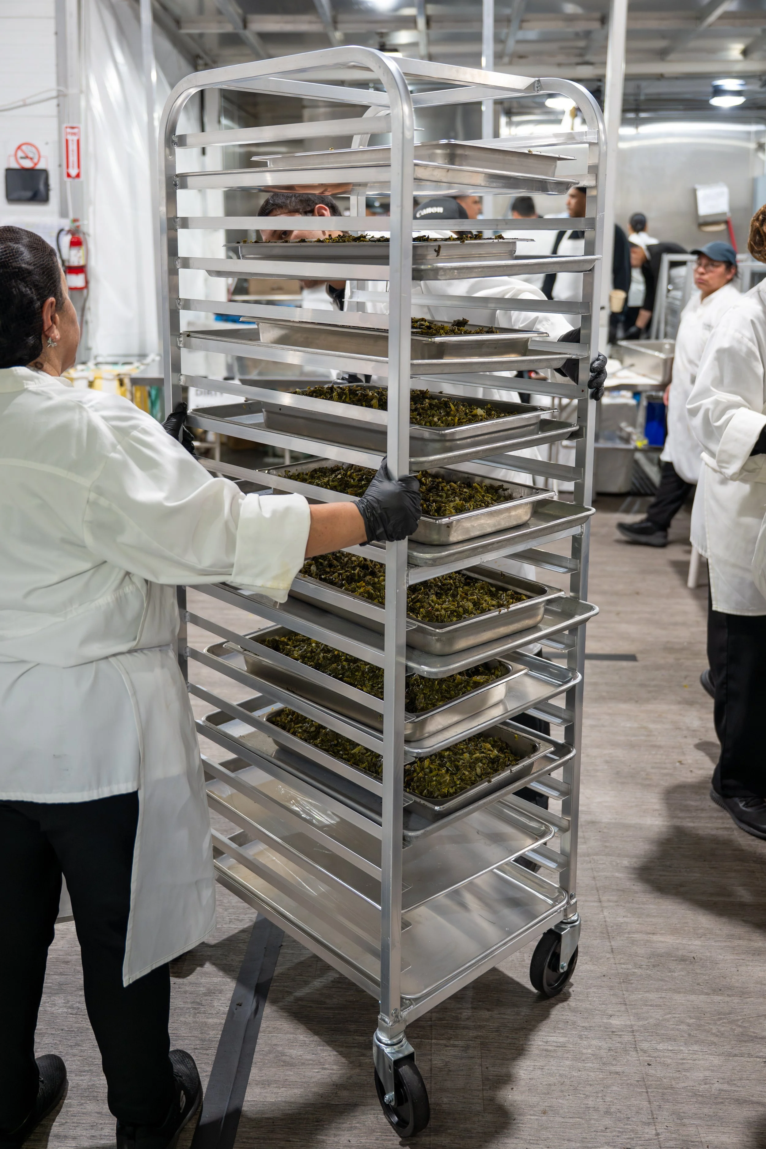 Kitchen staff working with trays of cooked leafy greens on a rolling metal rack in a commercial kitchen.