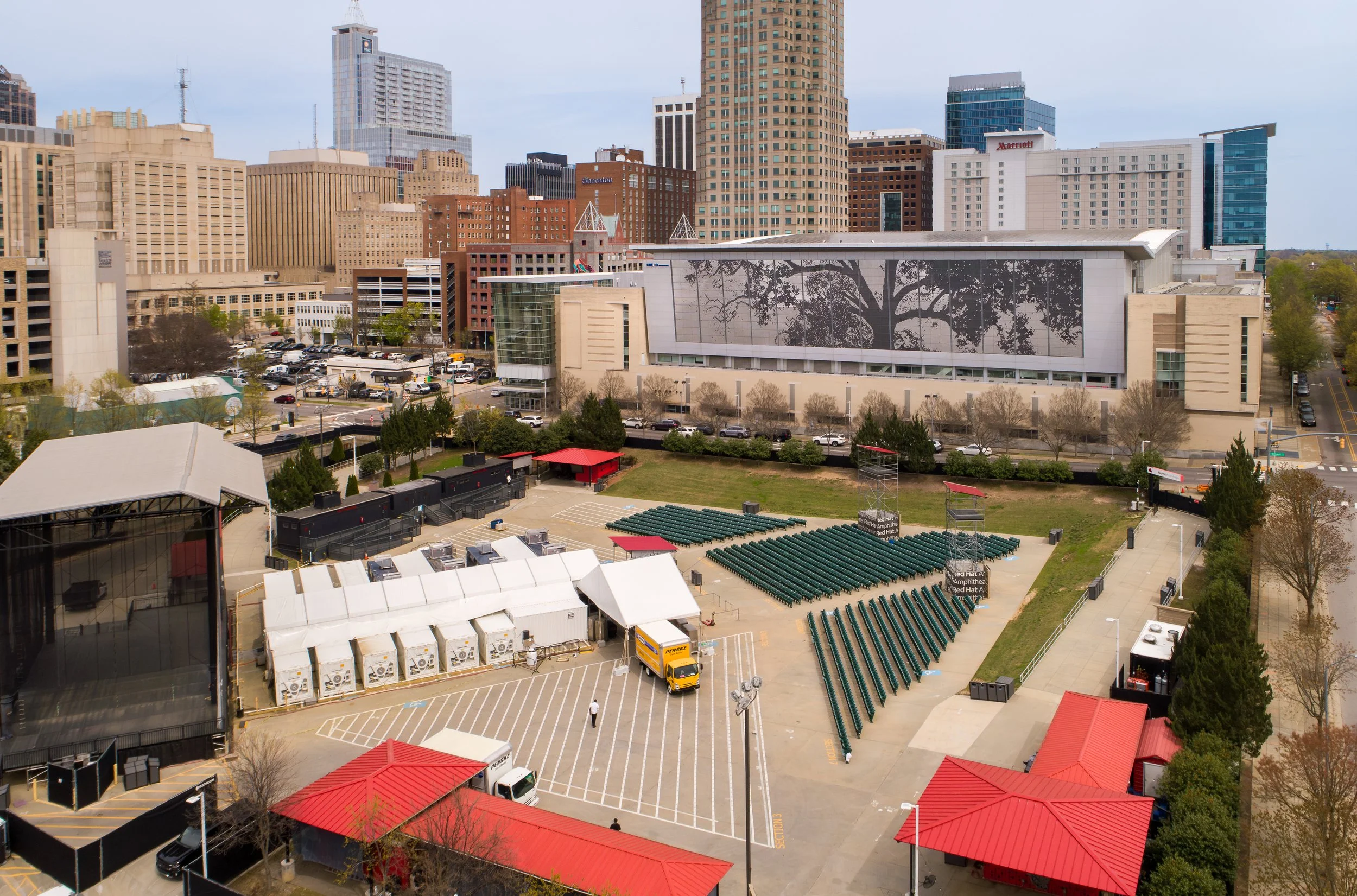 City skyline with tall buildings in the background and a large arena or auditorium in the foreground, equipped with seating and tents, and a parking lot with vehicles.