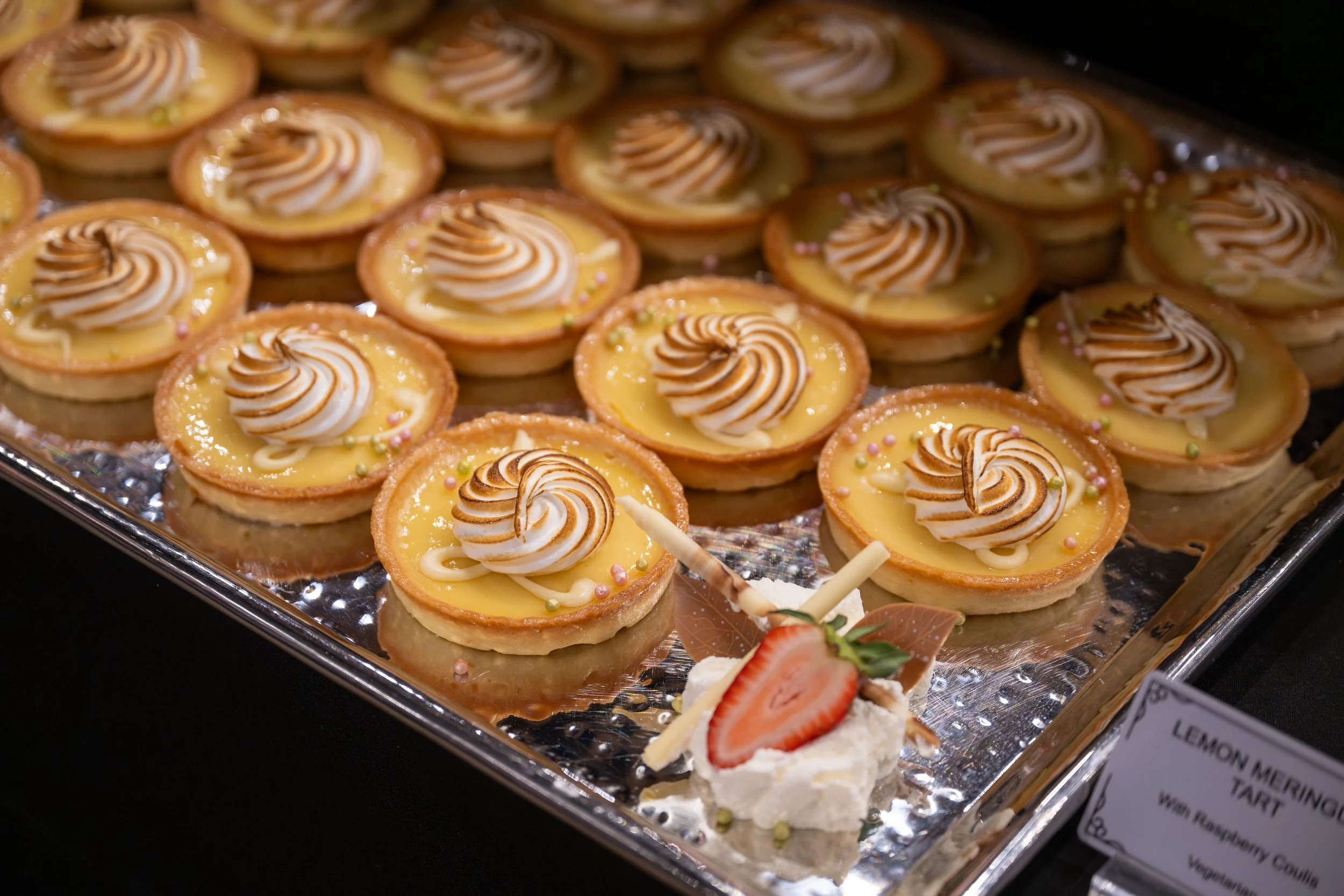 Tray of lemon meringue tartlets topped with toasted meringue swirl, decorated with small pastel-colored sugar pearls, and accompanied by a strawberry dessert with whipped cream and strawberry slices.