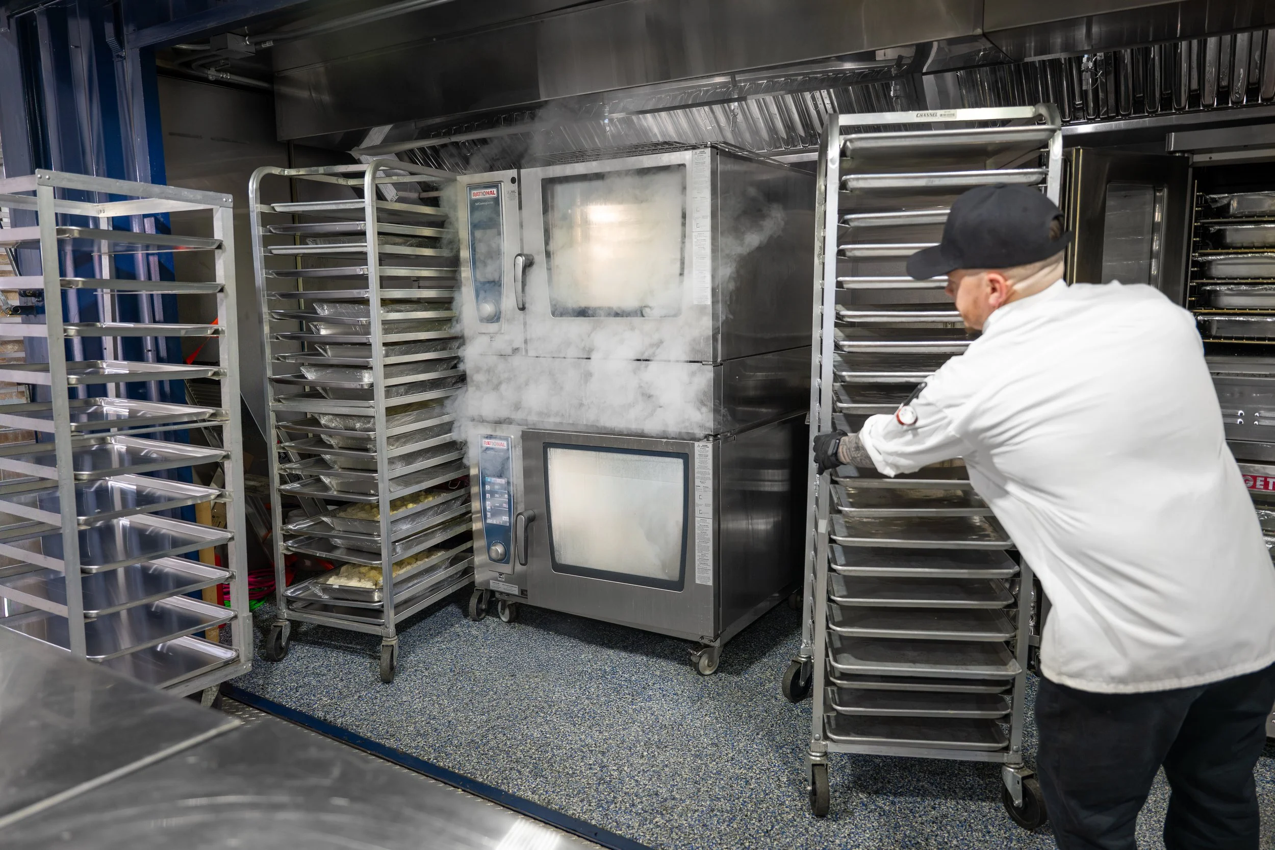 A person wearing a white coat and black cap arranging trays in a commercial kitchen with stainless steel equipment and racks.