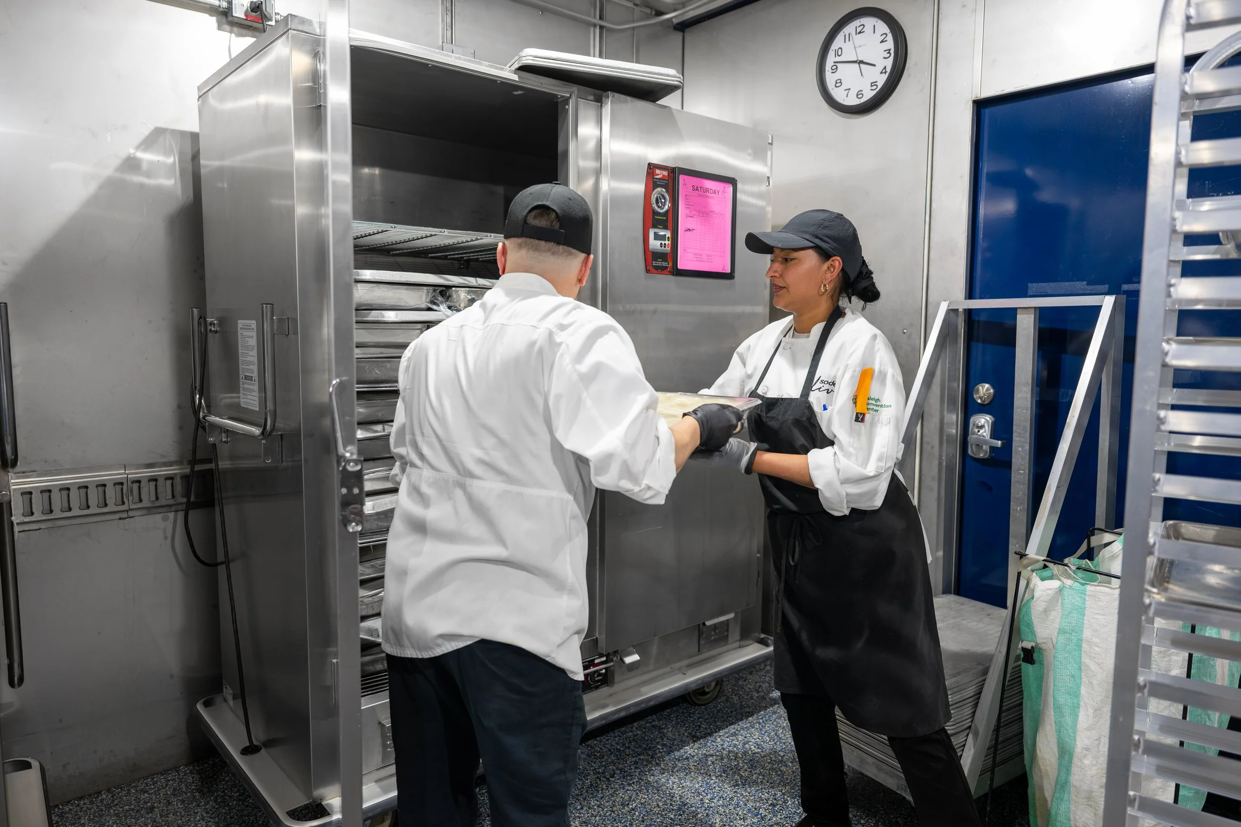 Two workers in a commercial kitchen or bakery, wearing white coats and black caps, loading a large hotbox with trays of food.