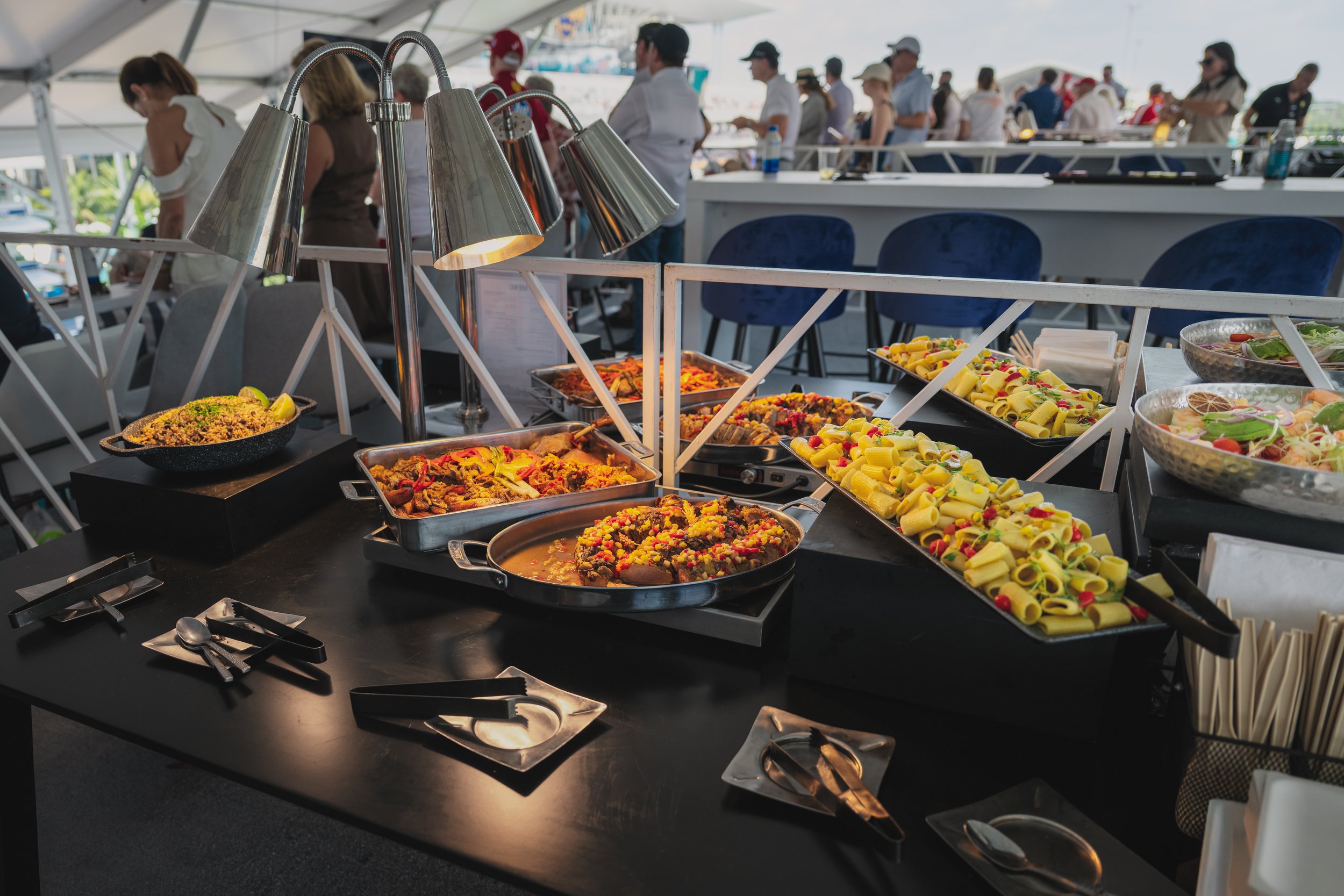 Buffet table with various dishes including pasta salad, chili, and salads at an outdoor event with people in the background.